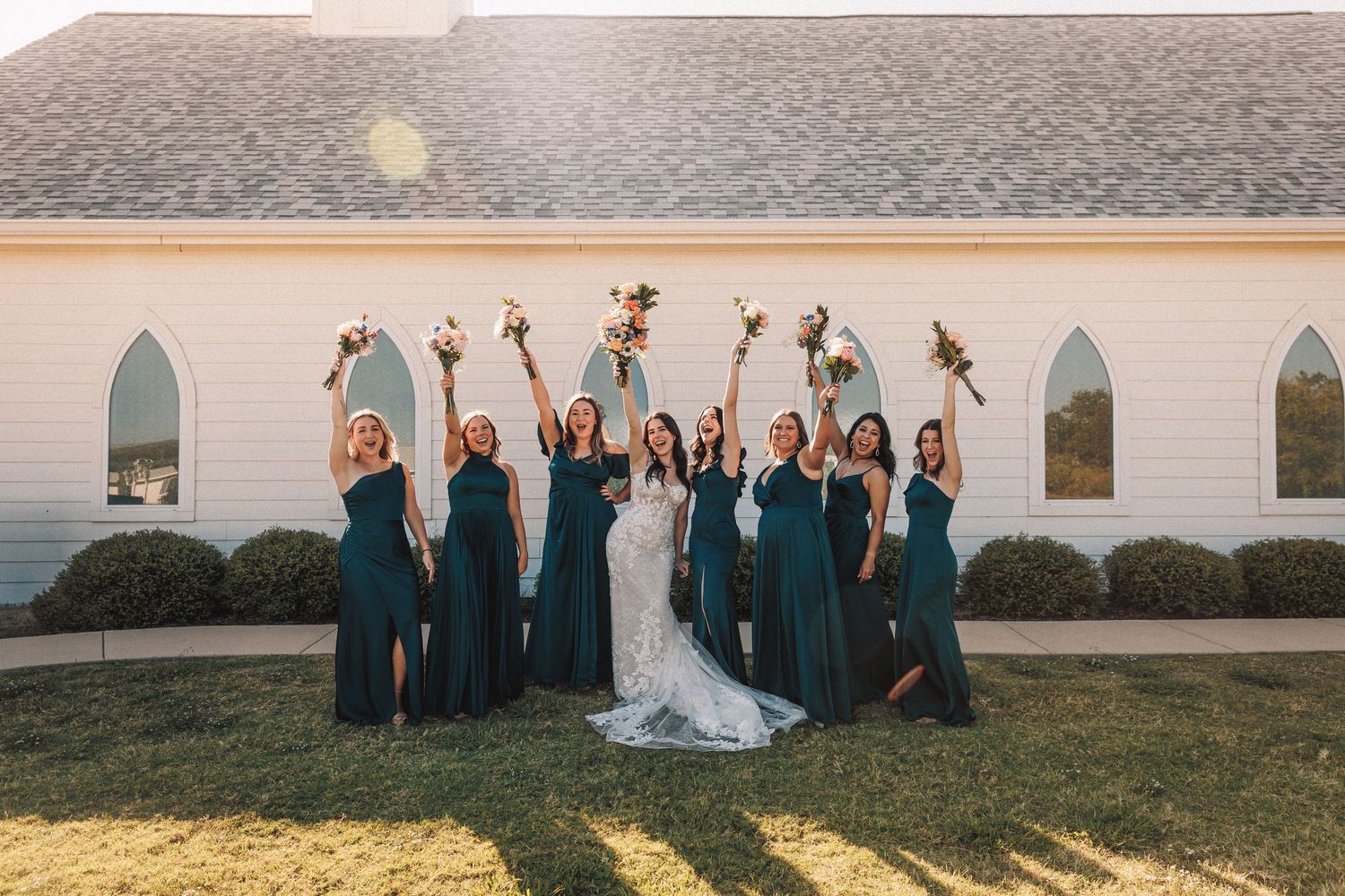 Bride with bridesmaids in emerald green gowns raising bouquets outside white chapel wedding venue.
