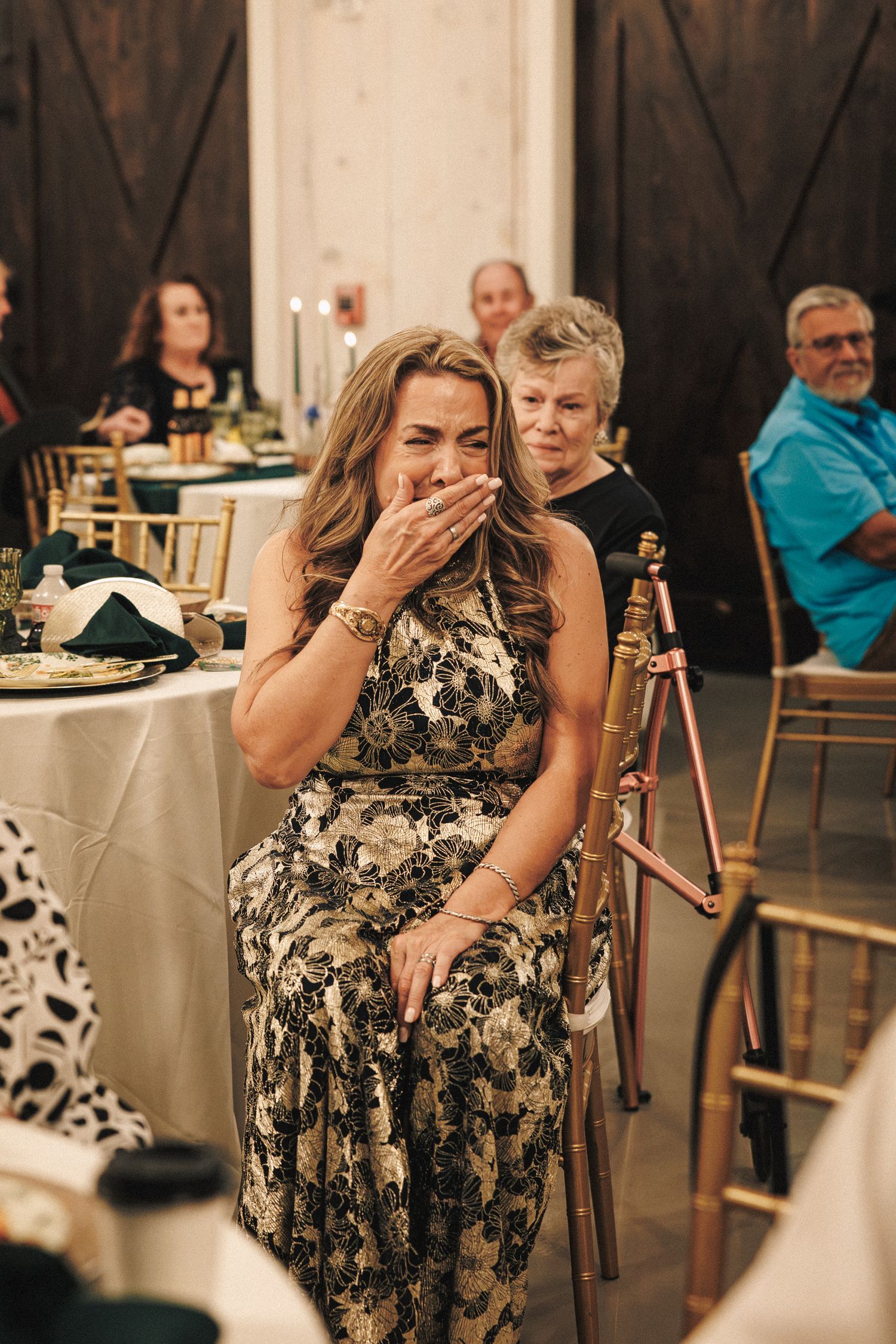 Emotional wedding guest covers her mouth in tears during heartfelt reception speech at barn venue.