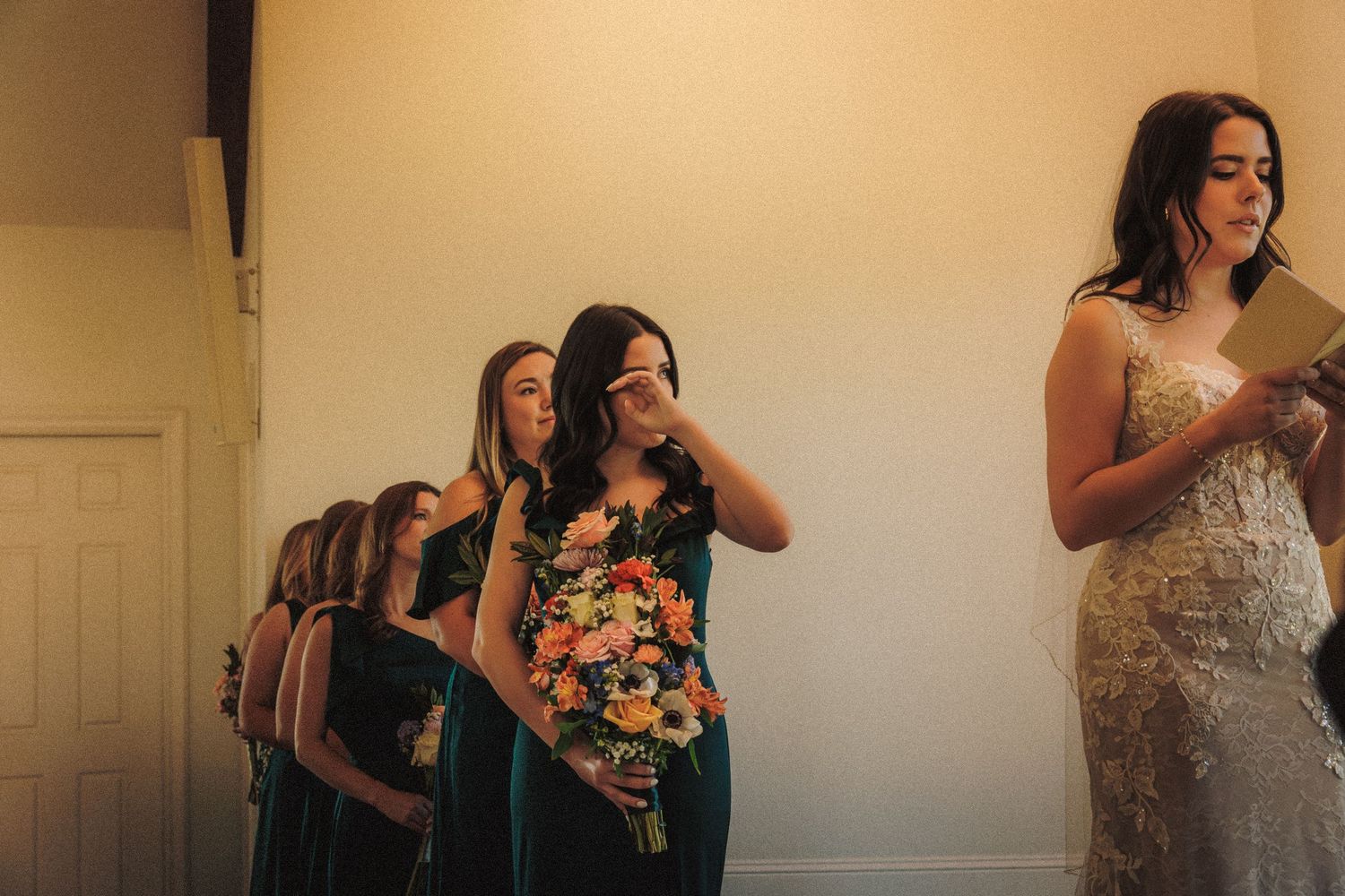 Bridesmaids in green dresses holding bouquets while bride reads vows, one wiping tears during wedding ceremony.