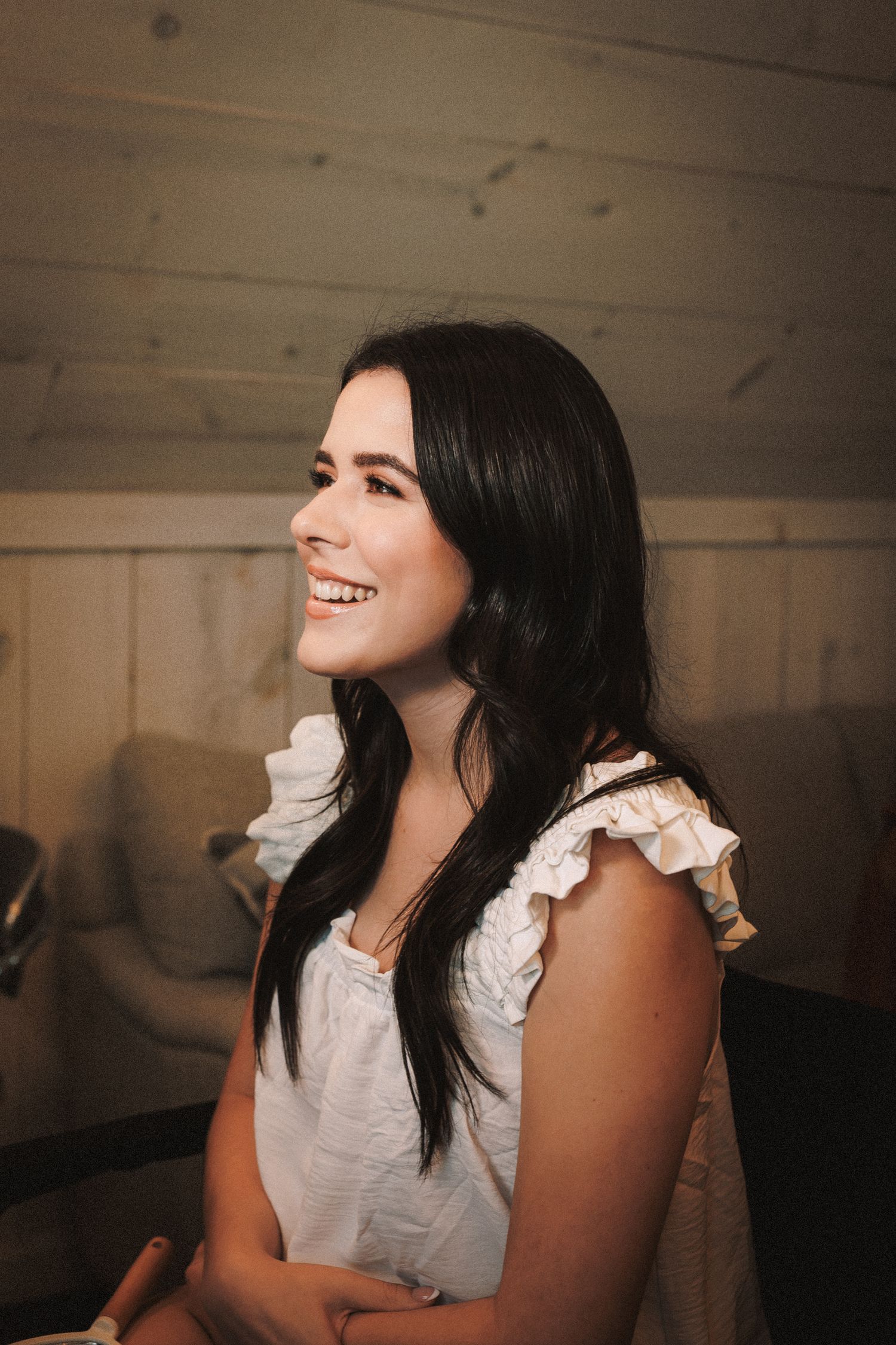 Smiling dark-haired bride in white ruffled top during wedding preparations indoors.