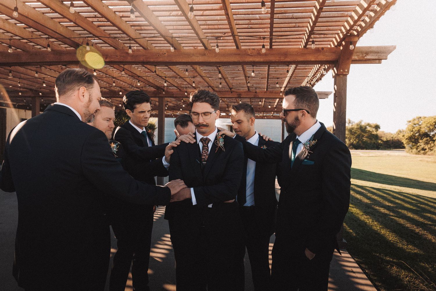 Groomsmen in black suits shaking hands and gathering under a wooden pergola at a wedding.