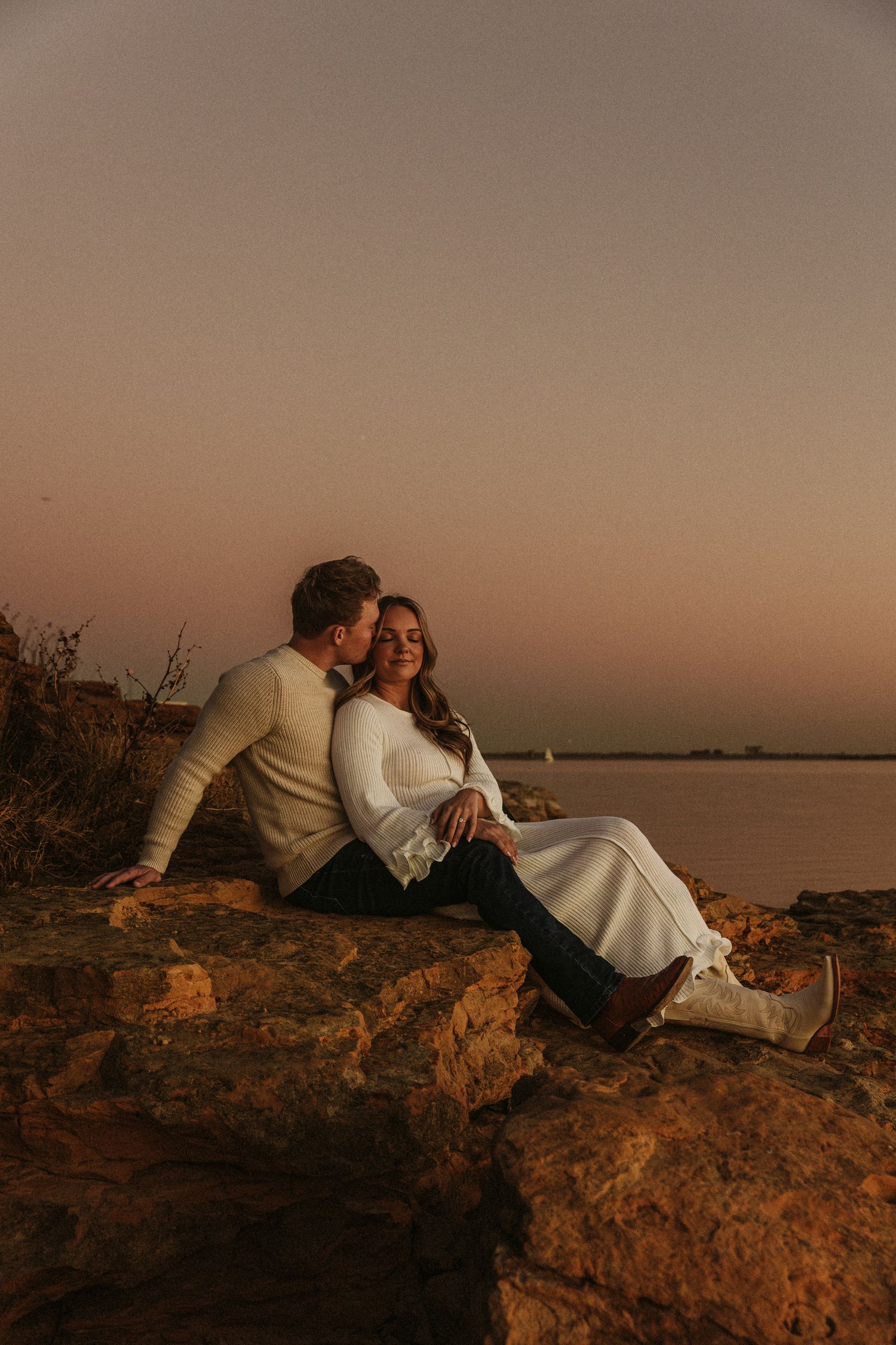 A couple reclines on coastal rocks during sunset in warm evening light.