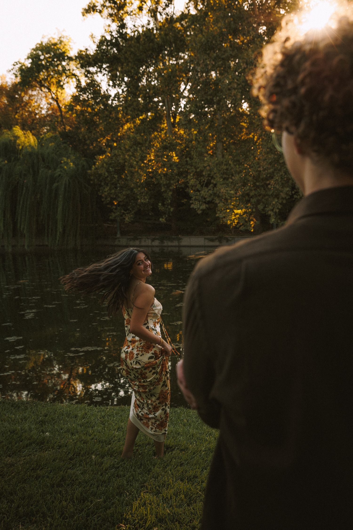 Two people share a playful moment outdoors near a pond during golden hour.