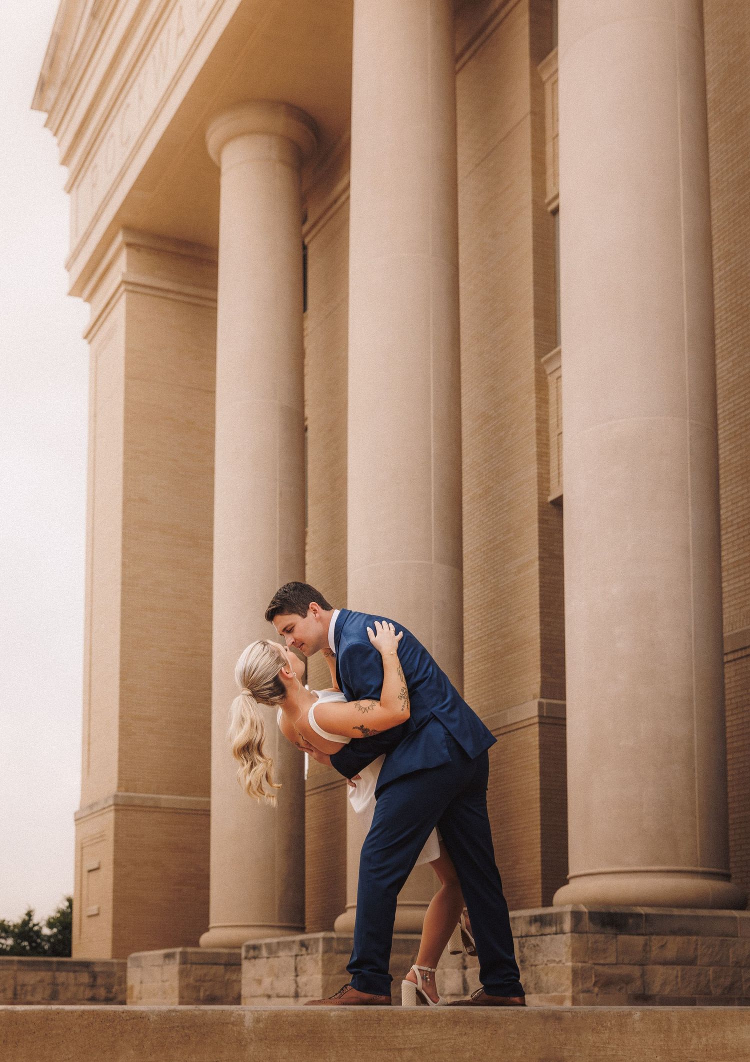 Dancers in navy blue outfits perform graceful movements between classical stone columns at sunset.