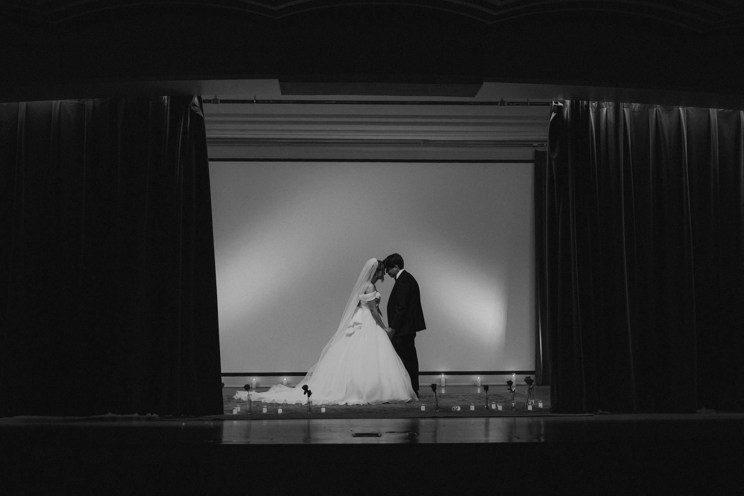 Bride and groom sharing romantic kiss on stage framed by dark curtains, black and white.