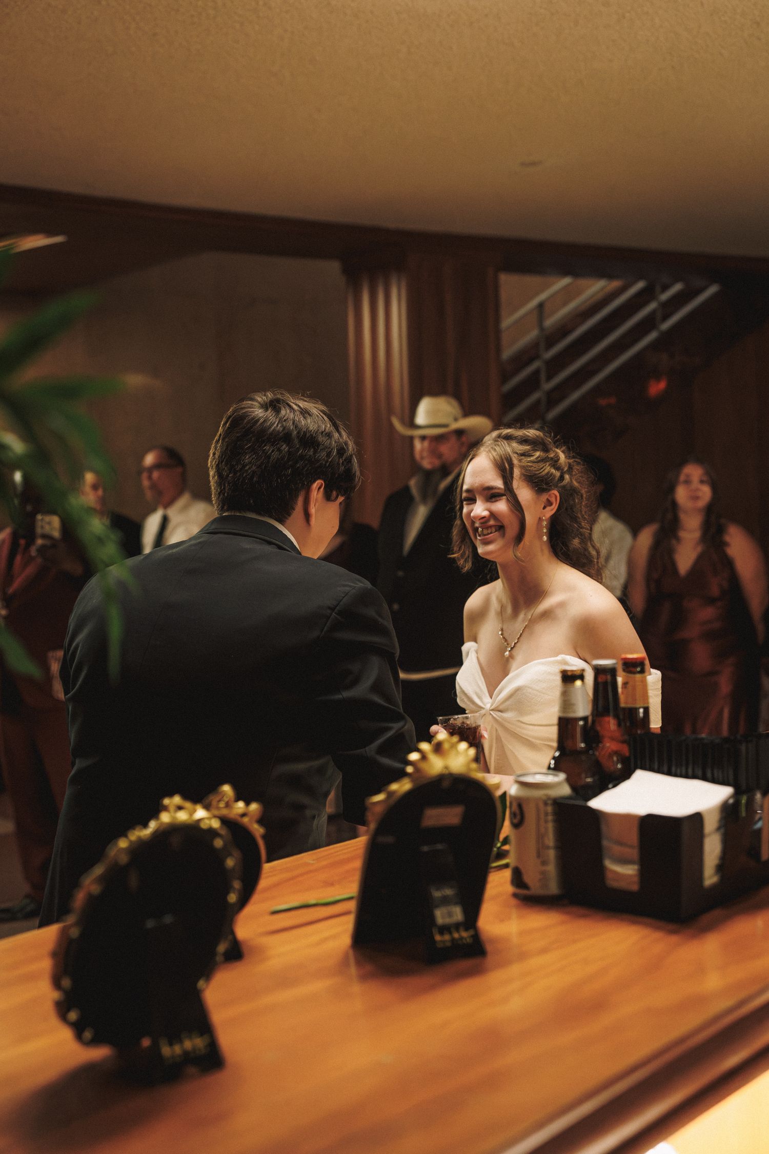 Smiling bride in white gown chatting with guest at a warmly lit wedding reception bar.