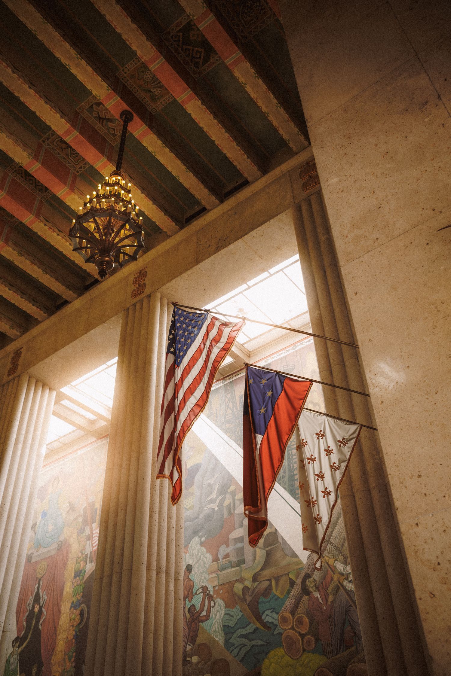 Sunlit grand hall interior with American flags, ornate columns, chandelier, and colorful murals.