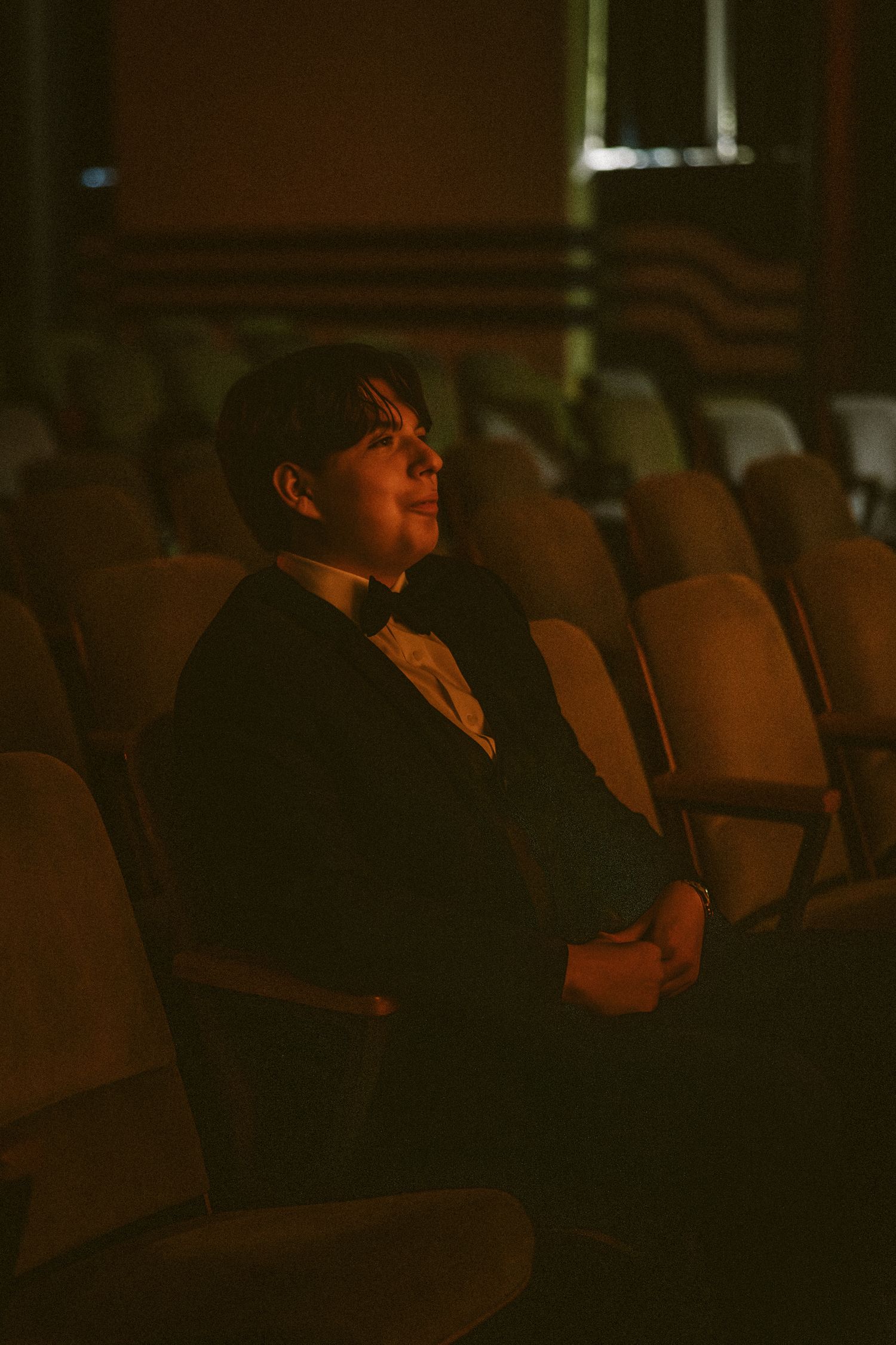 Young man in tuxedo sitting alone in dimly lit theater seats with warm amber lighting.
