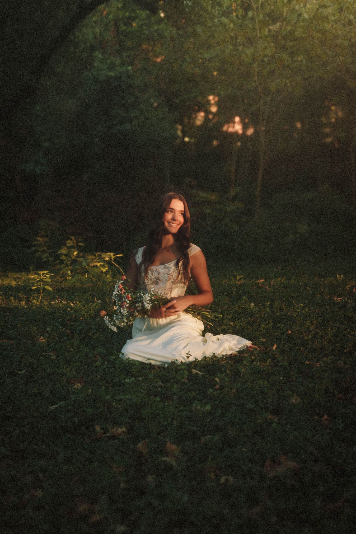 A person in a white dress sits in a sunlit forest clearing at golden hour holding a bouquet of wildflowers.