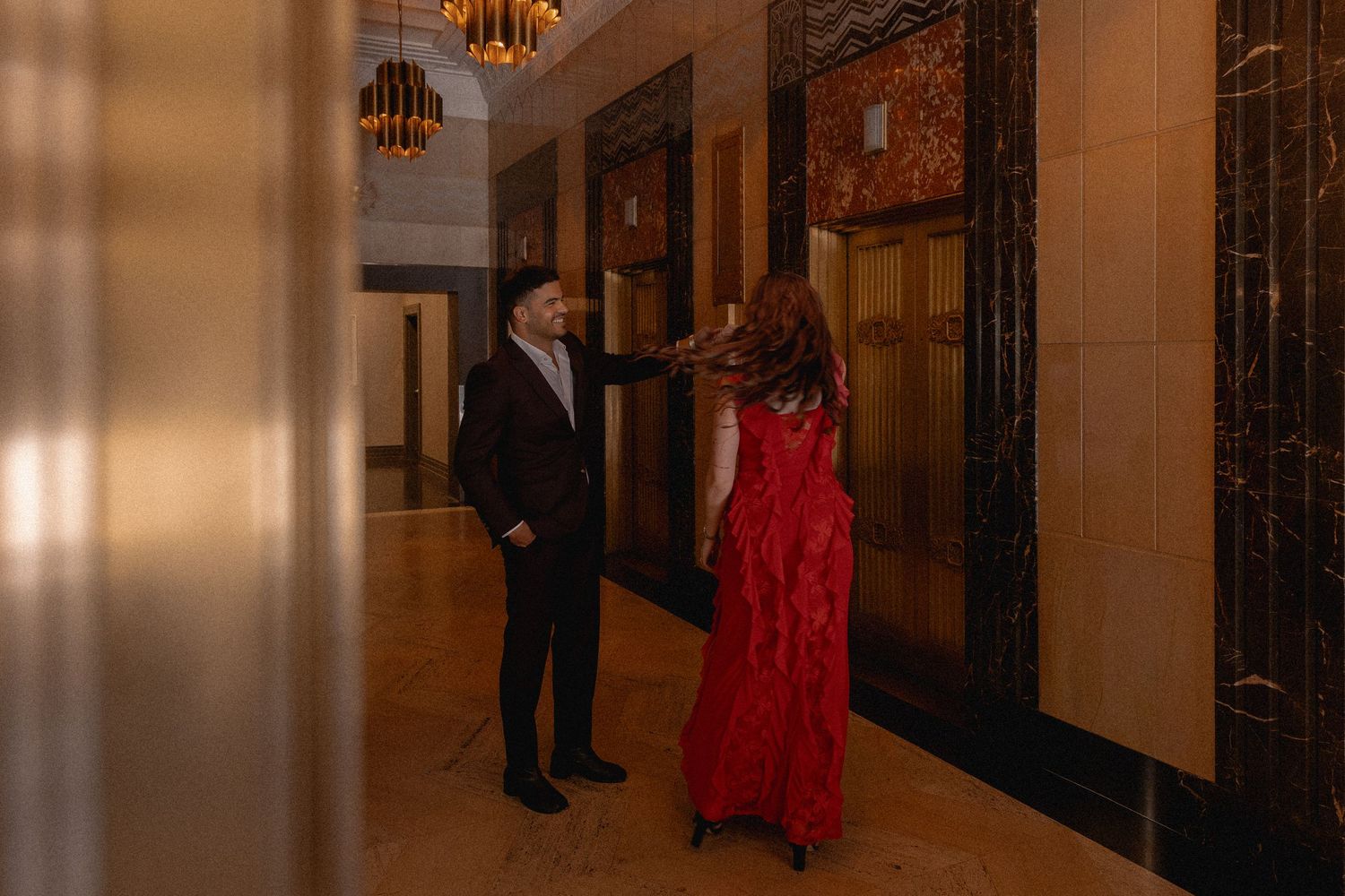 Couple in elegant attire, man in suit and woman in red gown, conversing in a luxurious marble hallway.