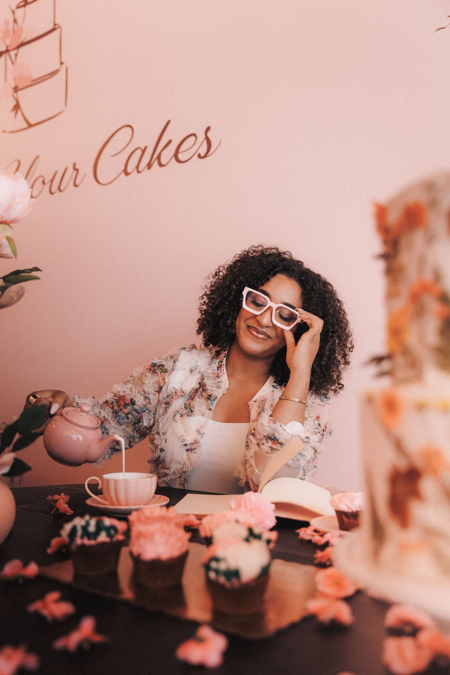 Person with curly hair and glasses enjoys tea time with floral decor and soft pink lighting in styled photoshoot.
