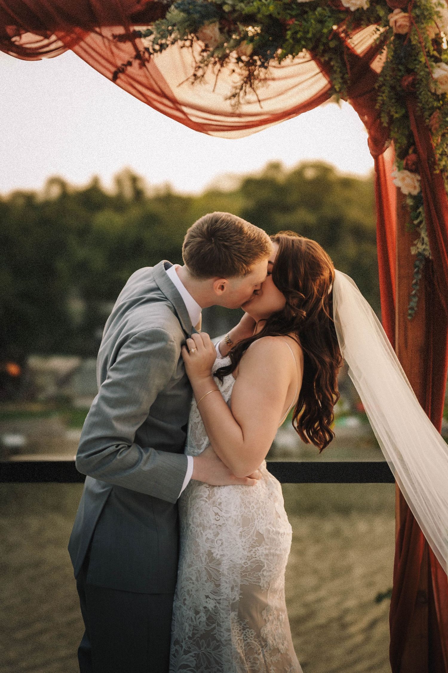 Romantic wedding couple shares intimate moment under wooden arch draped with fabric at sunset.