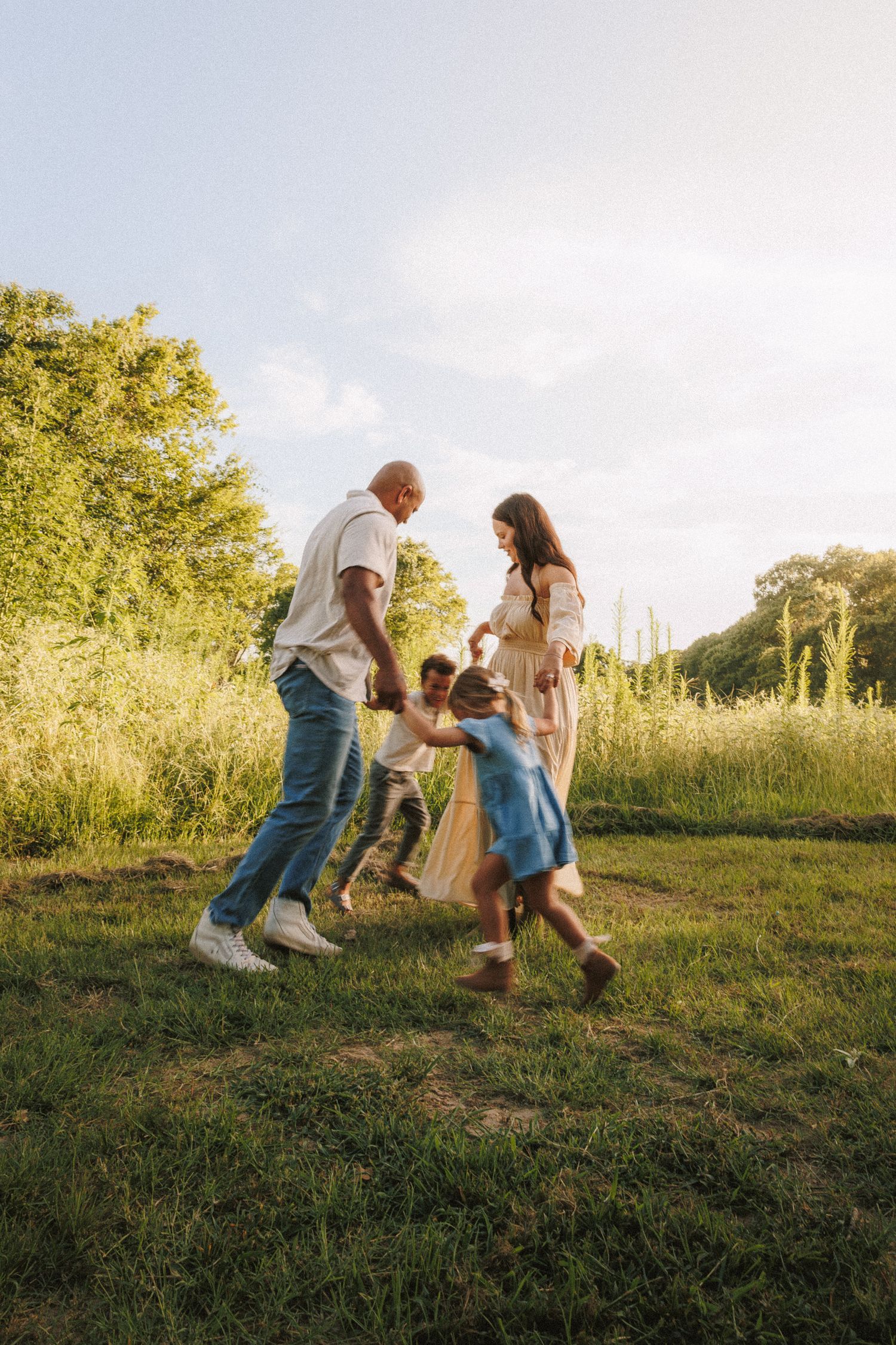Family playing and dancing together in a sunny meadow with trees in the background during golden hour.