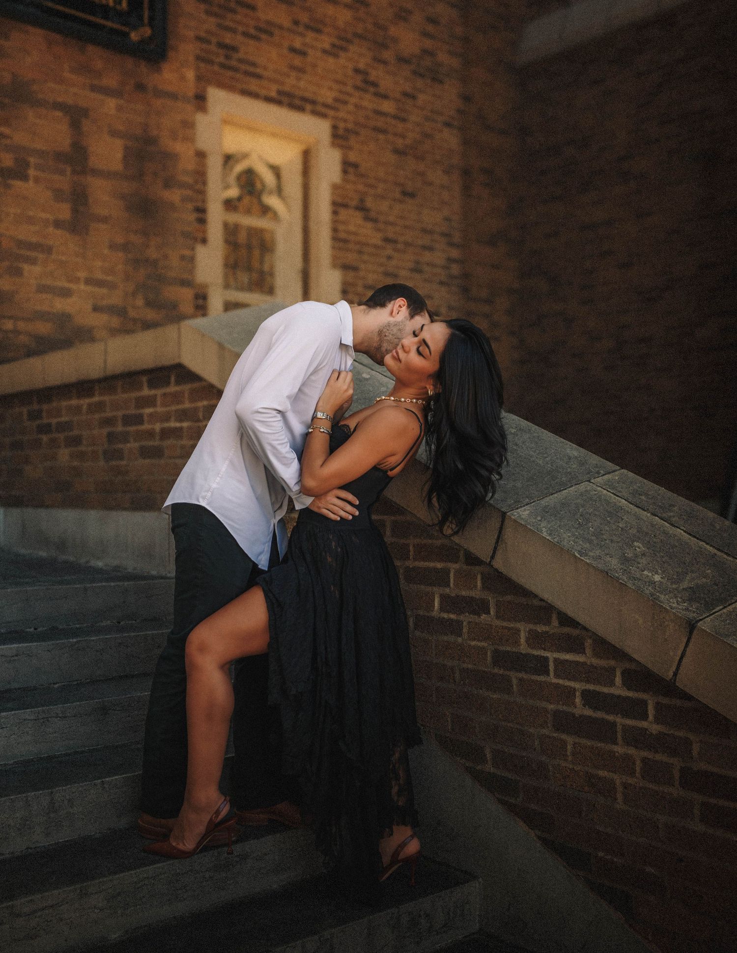 A couple shares an intimate moment on stone steps against a rustic brick wall in warm lighting.