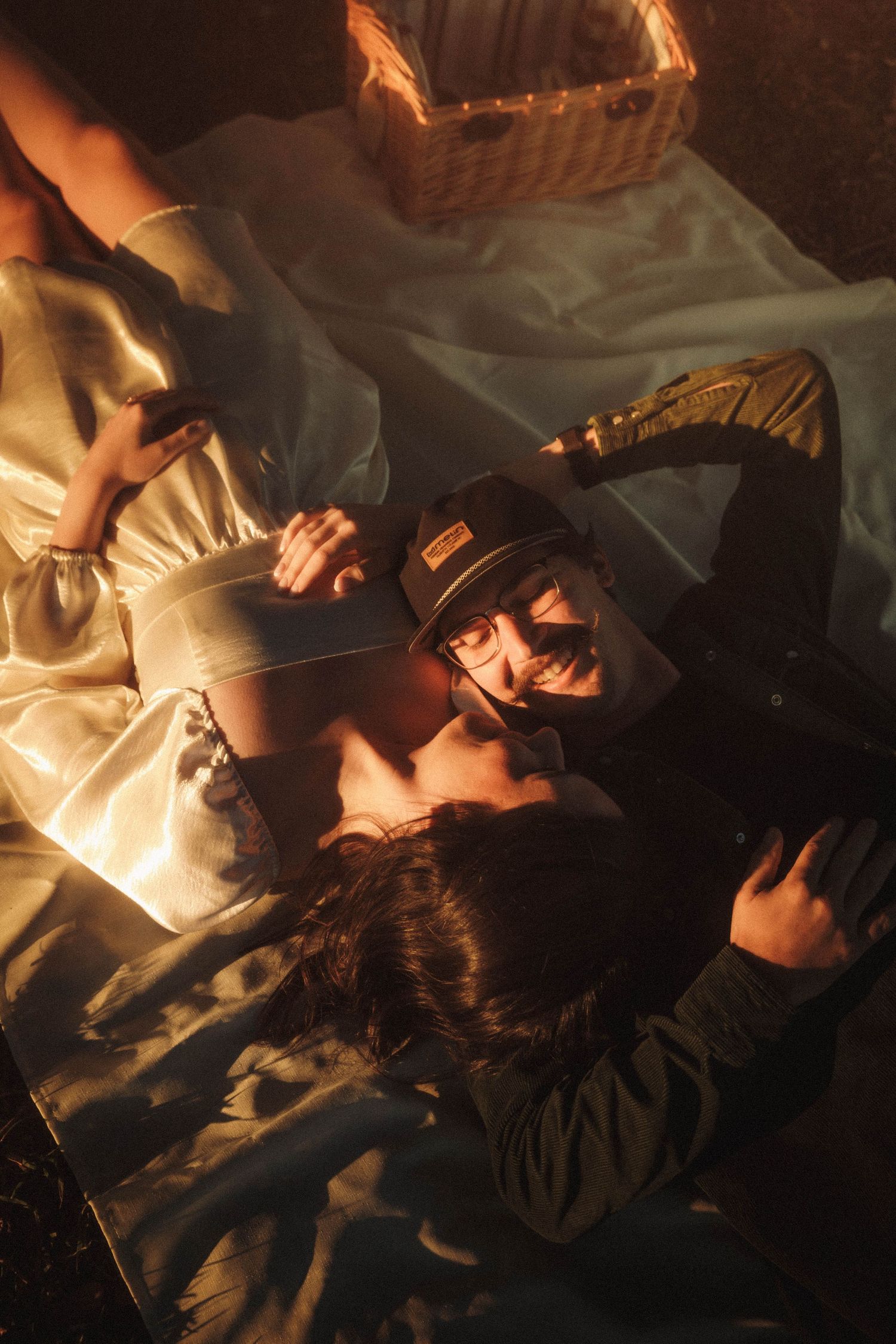 Atmospheric photo taken in warm lighting shows someone laying down beside a wicker basket on white fabric.