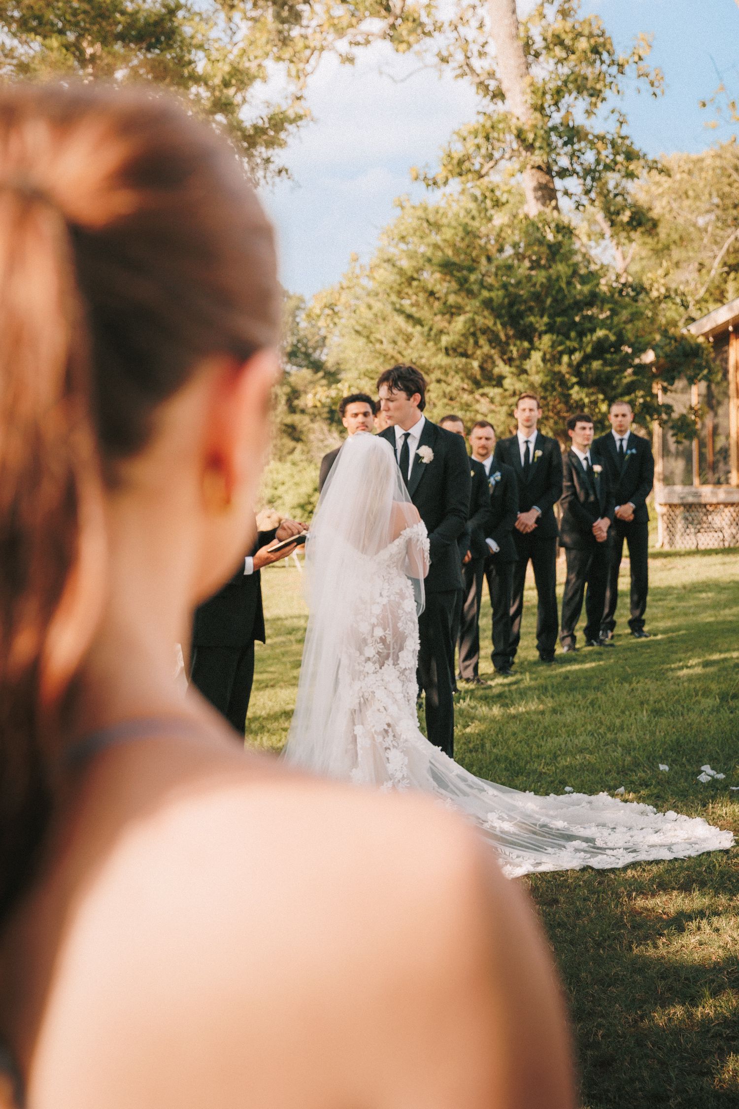 Wedding party in formal attire stands in a line on grassy lawn during autumn afternoon ceremony.