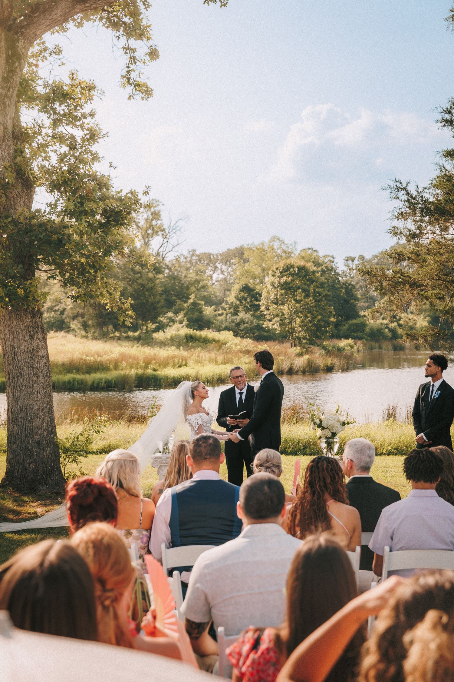 Outdoor wedding ceremony with guests seated in white chairs overlooking a serene lake surrounded by autumn trees.