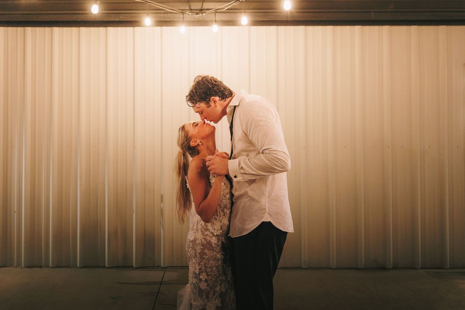 Romantic moment under warm lighting at wedding reception against white backdrop.