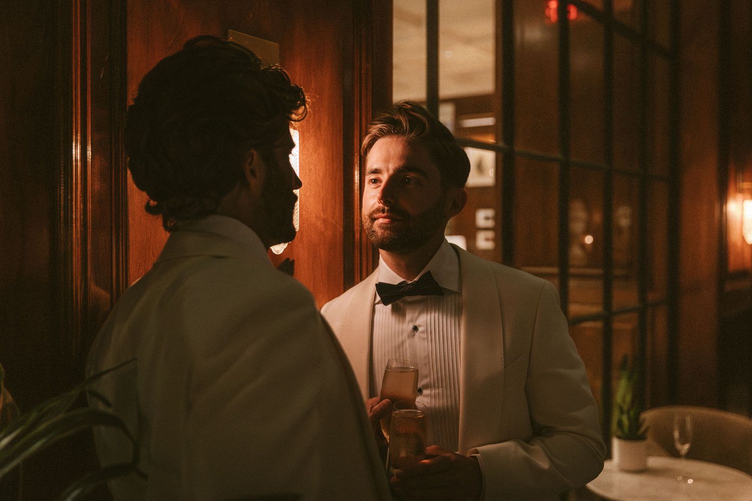 Two men in white tuxedos share a moment at an elegant candlelit wedding reception.