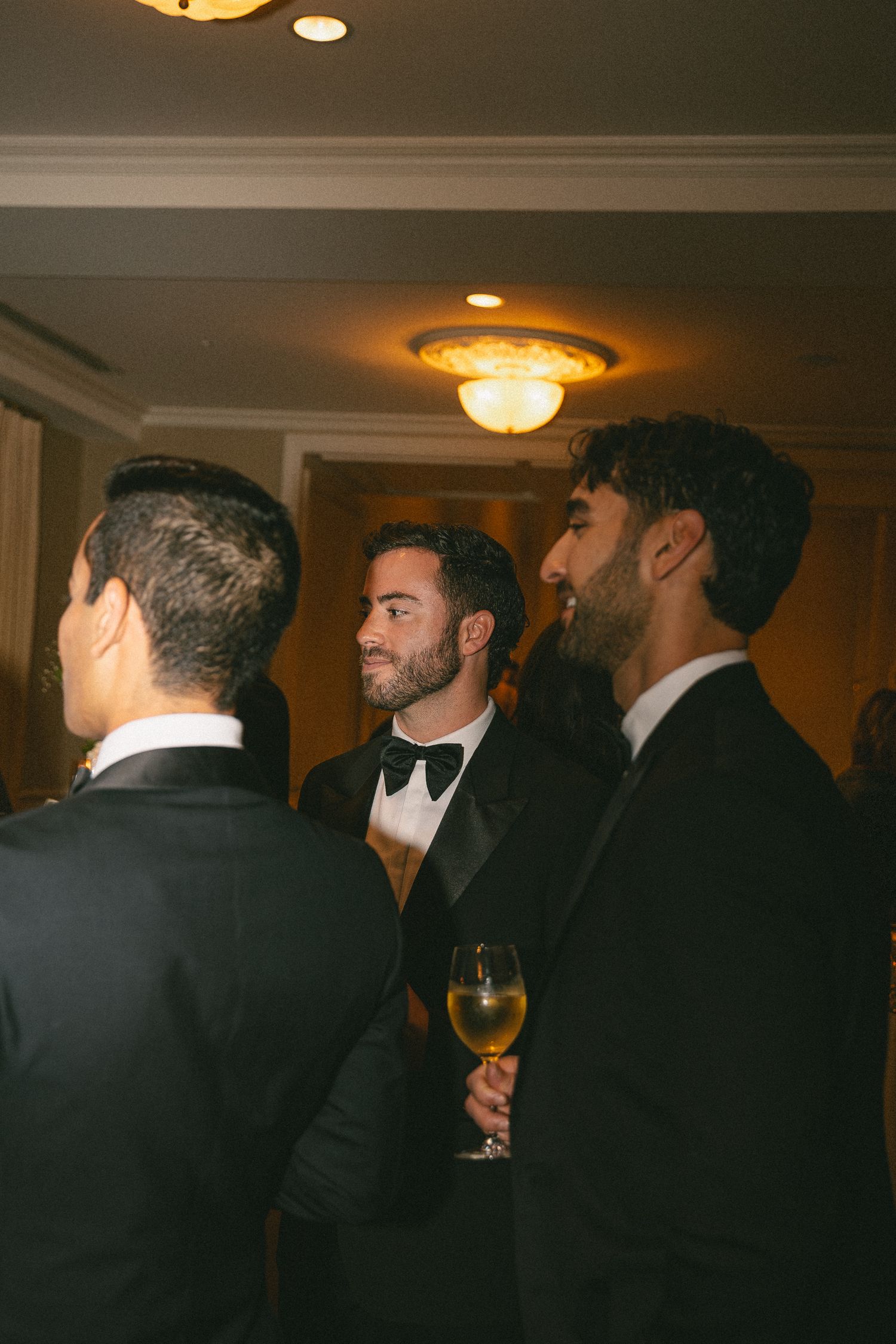 Guests in black tuxedos socializing at an elegant wedding reception with warm lighting.