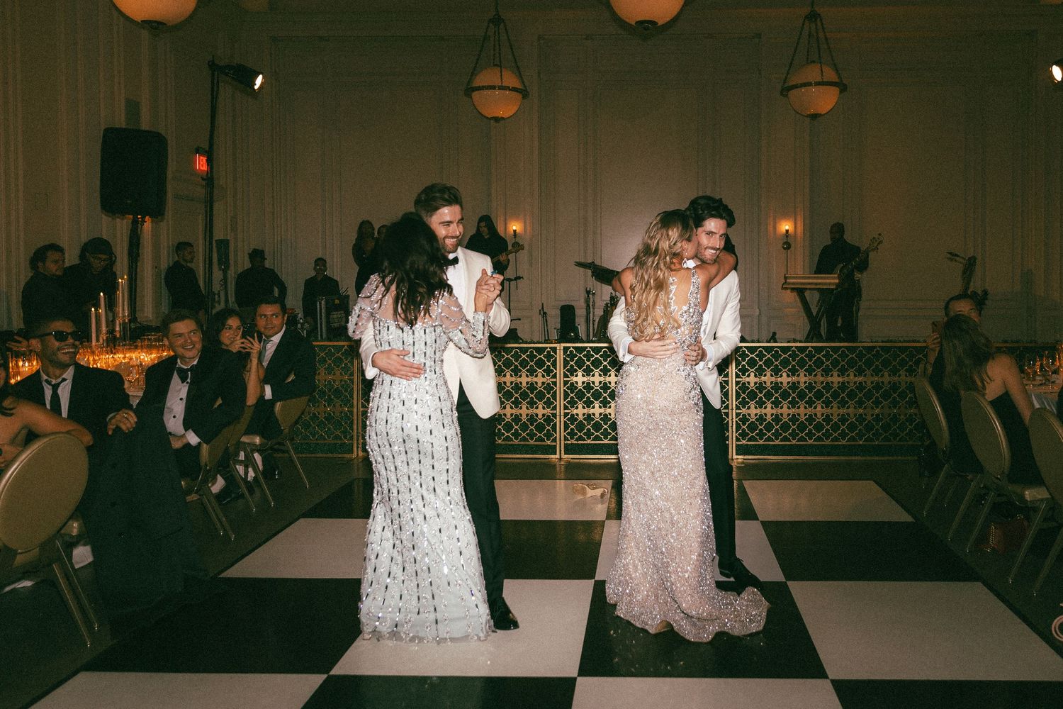 Two couples share their first dance on a black and white checkered floor at an elegant wedding reception.