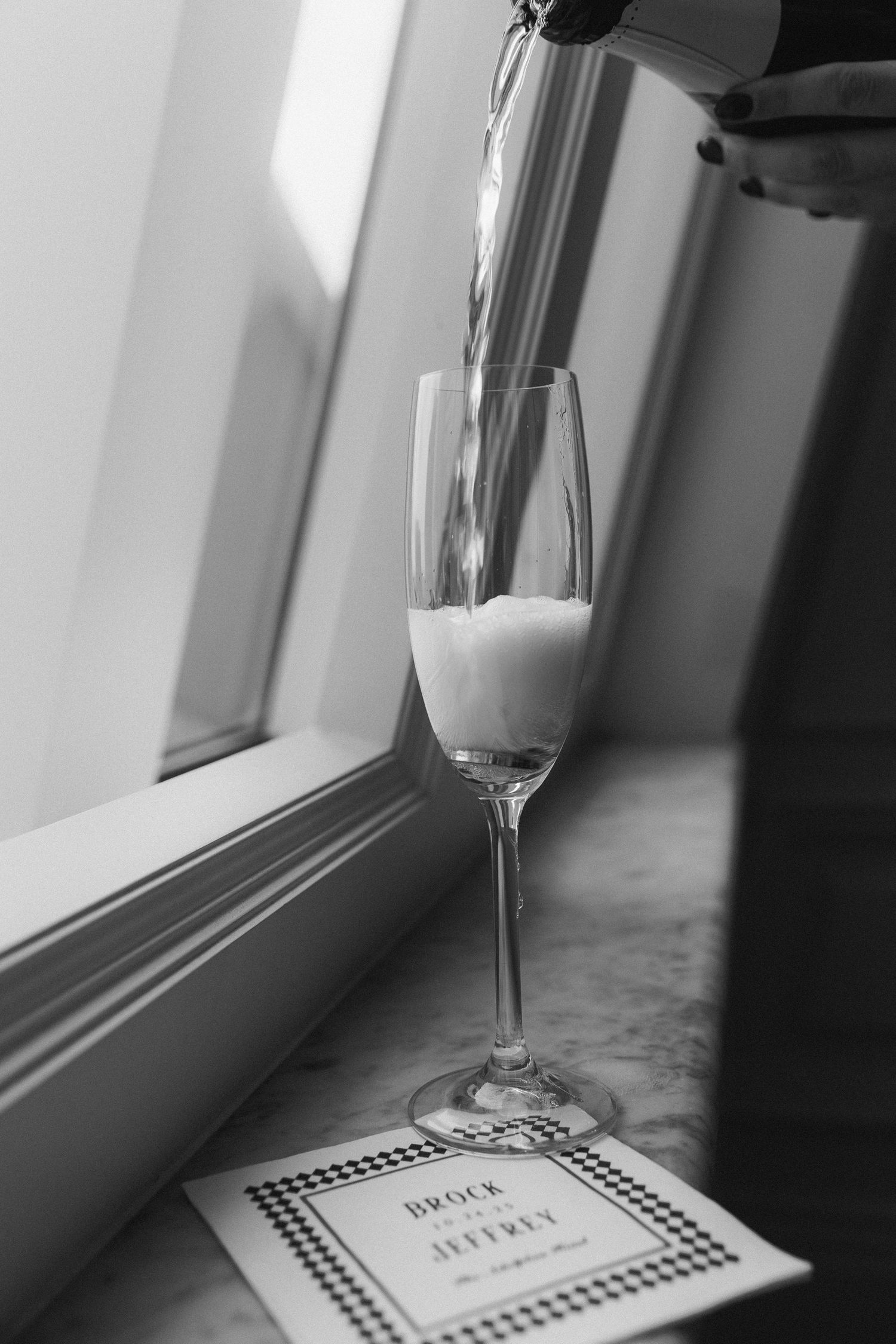 Champagne being poured into a flute beside a Brock and Jeffrey wedding program on a marble surface.
