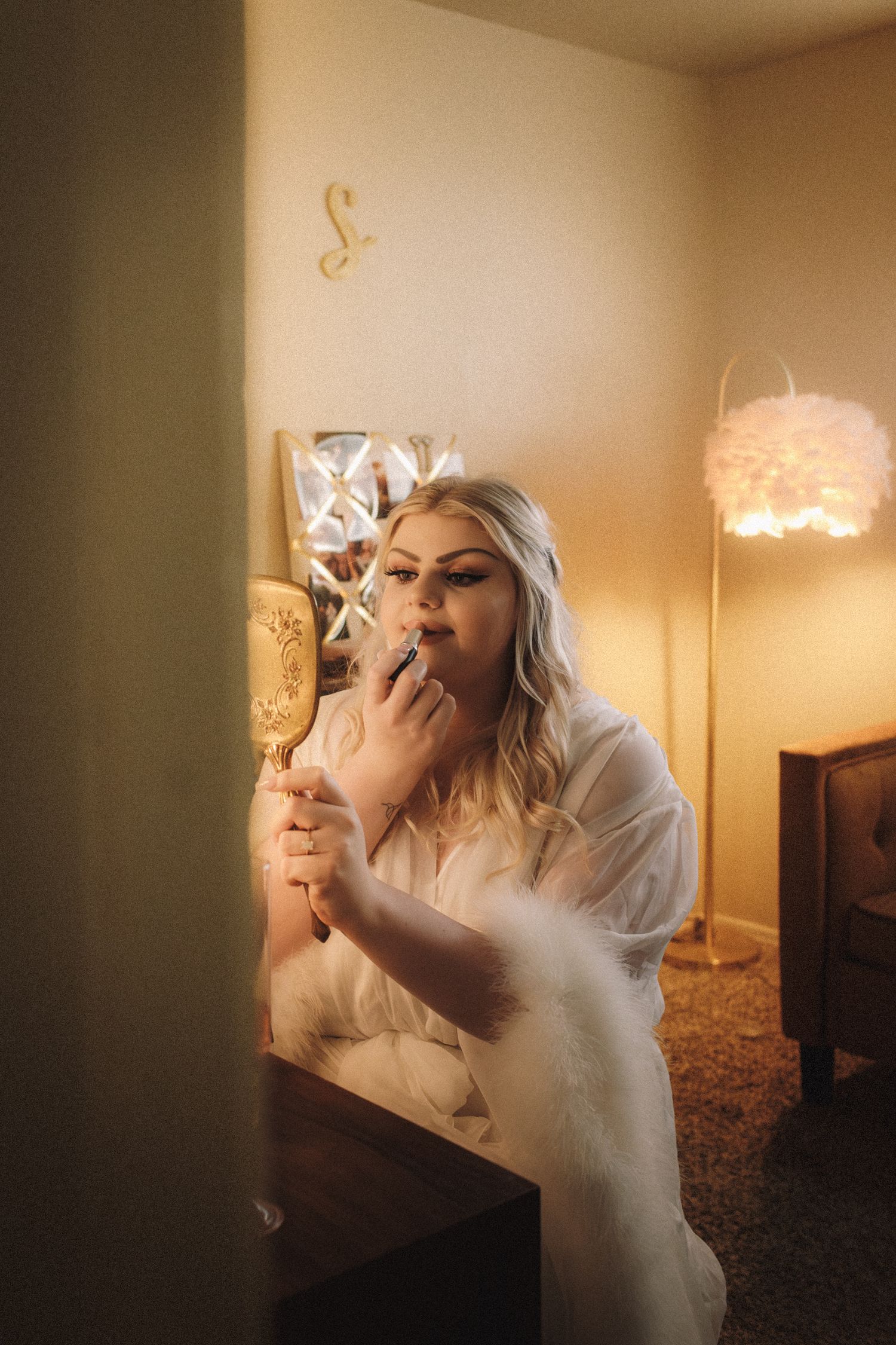 Bride in white fur-trimmed robe applying lipstick using ornate hand mirror in warm golden light.