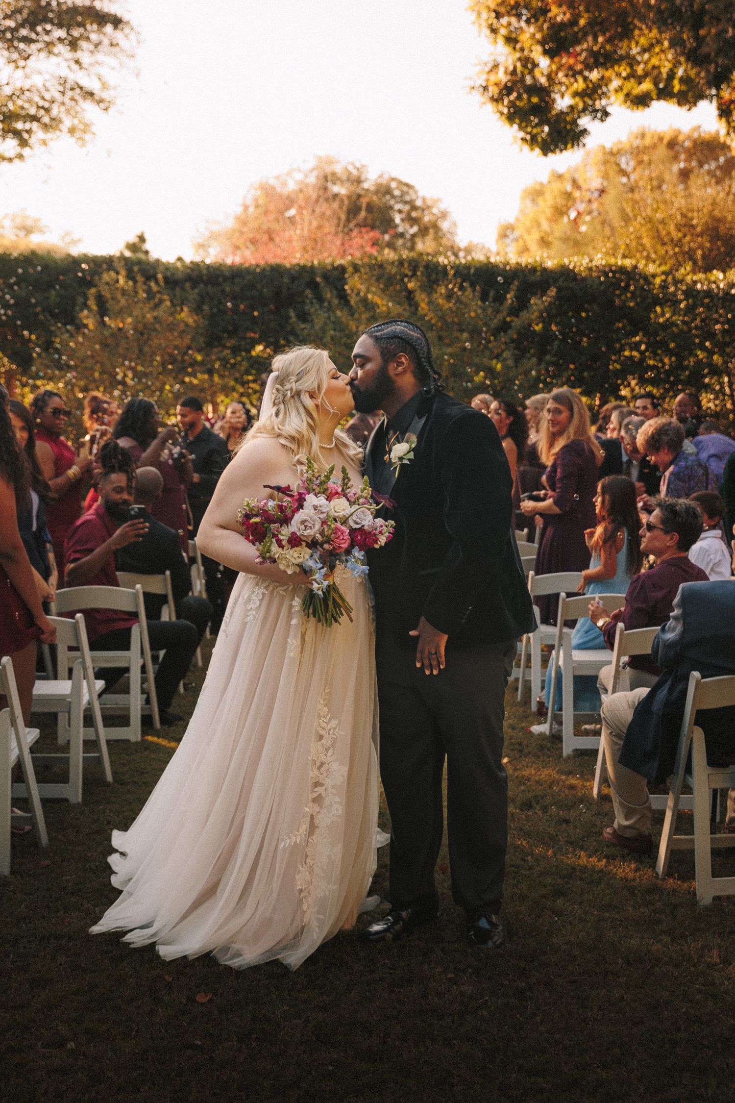 Newlywed couple shares a kiss at the altar during outdoor wedding ceremony with seated guests.