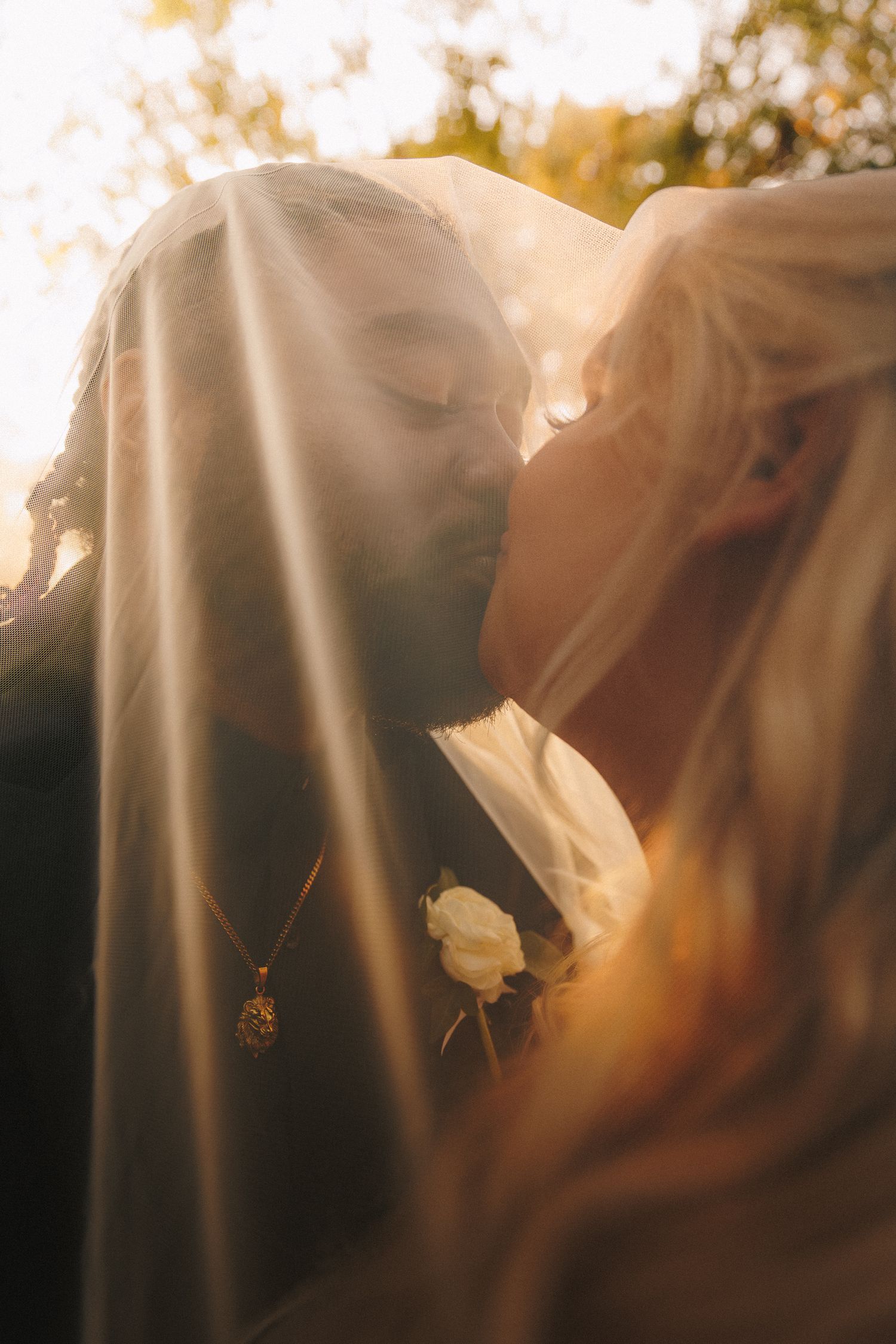 Romantic close-up of bride and groom kissing under veil in warm golden sunlight.