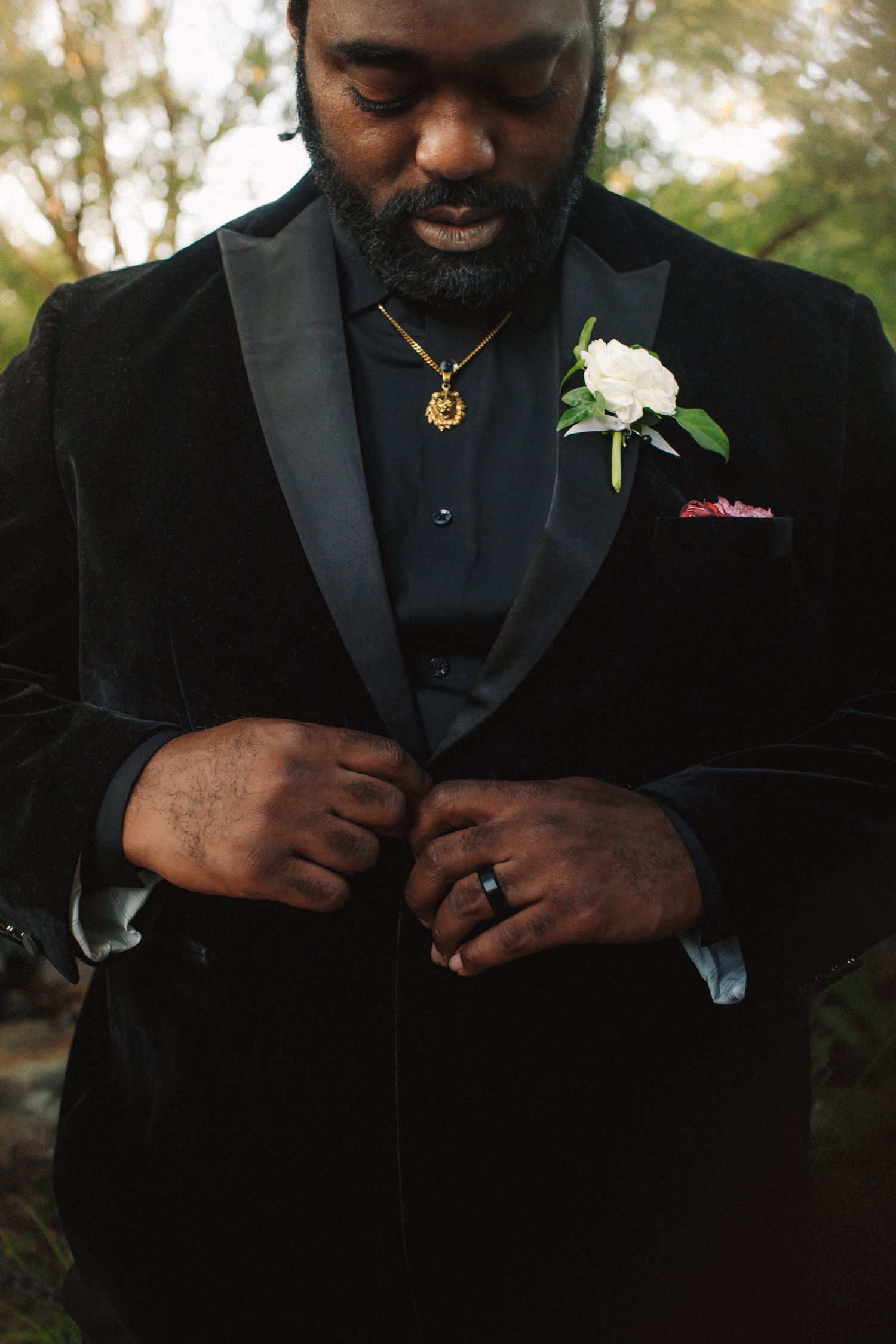 Groom in black velvet tuxedo with white rose boutonniere and gold chain necklace buttoning his jacket outdoors.