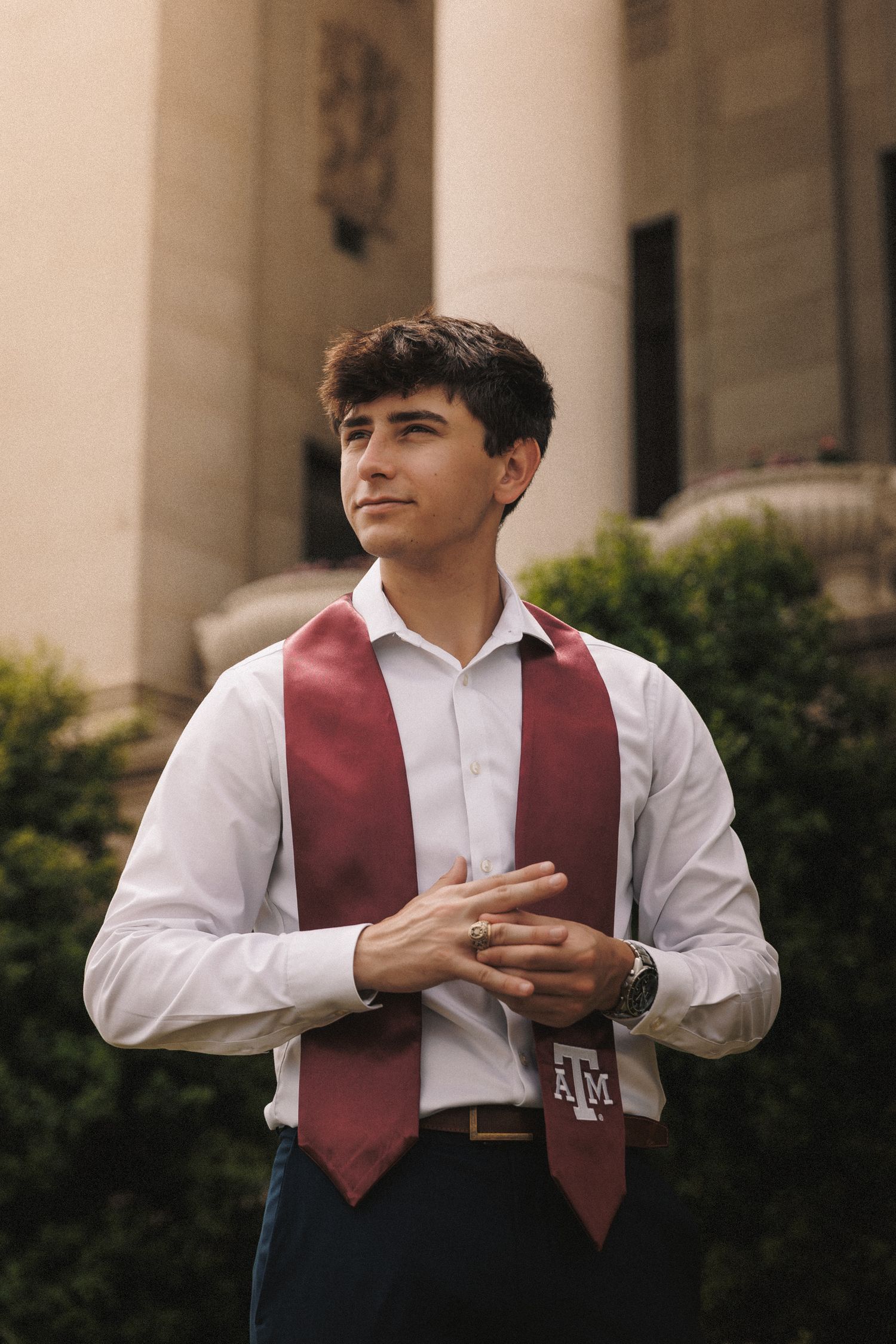 Young man in white shirt with maroon Texas A&M graduation stole poses outdoors near columns and greenery.