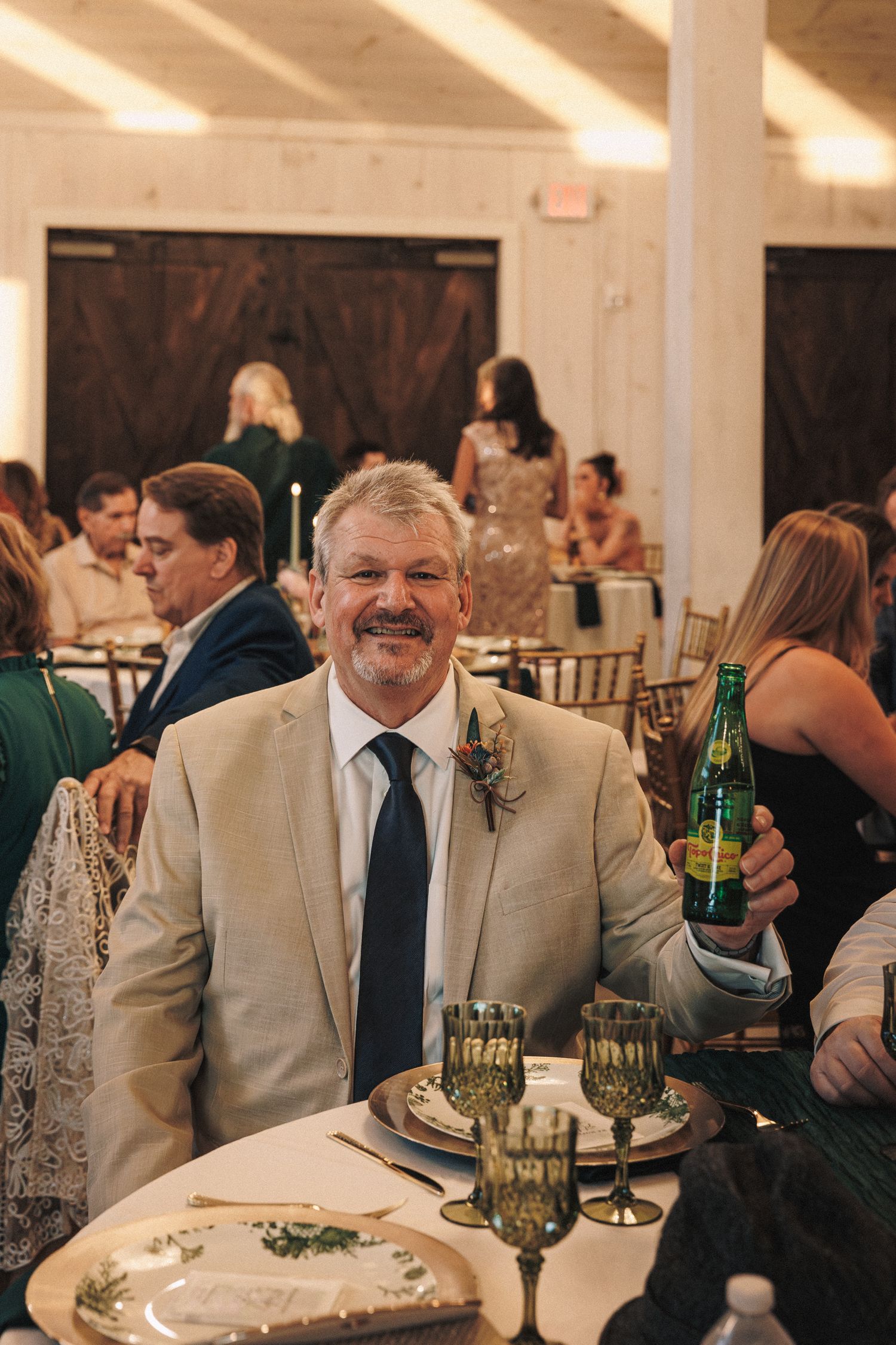 Smiling wedding guest in tan suit with boutonniere holds green bottle at elegantly decorated reception table.