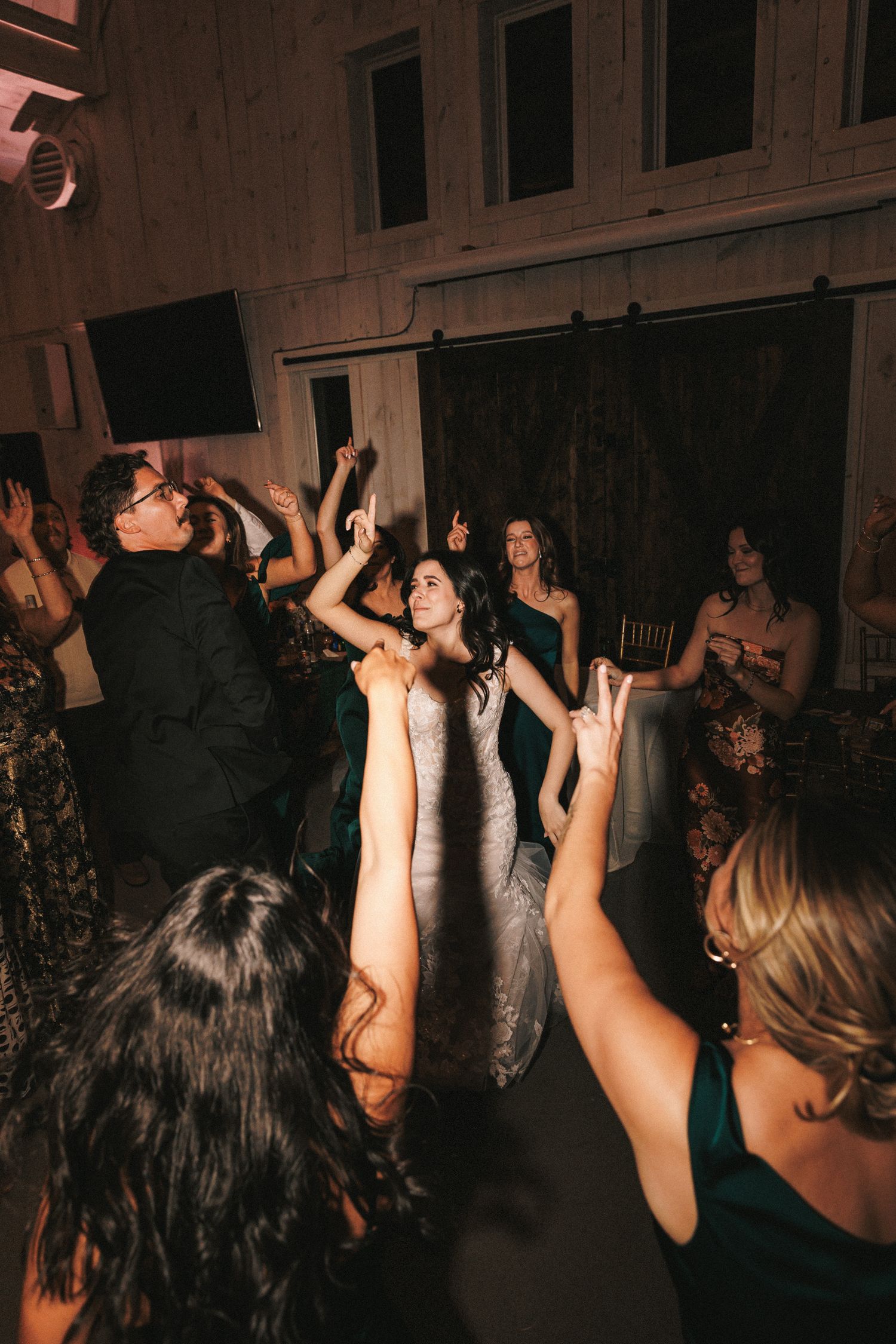 Bride in lace gown dances joyfully with wedding guests on crowded reception dance floor in barn venue.