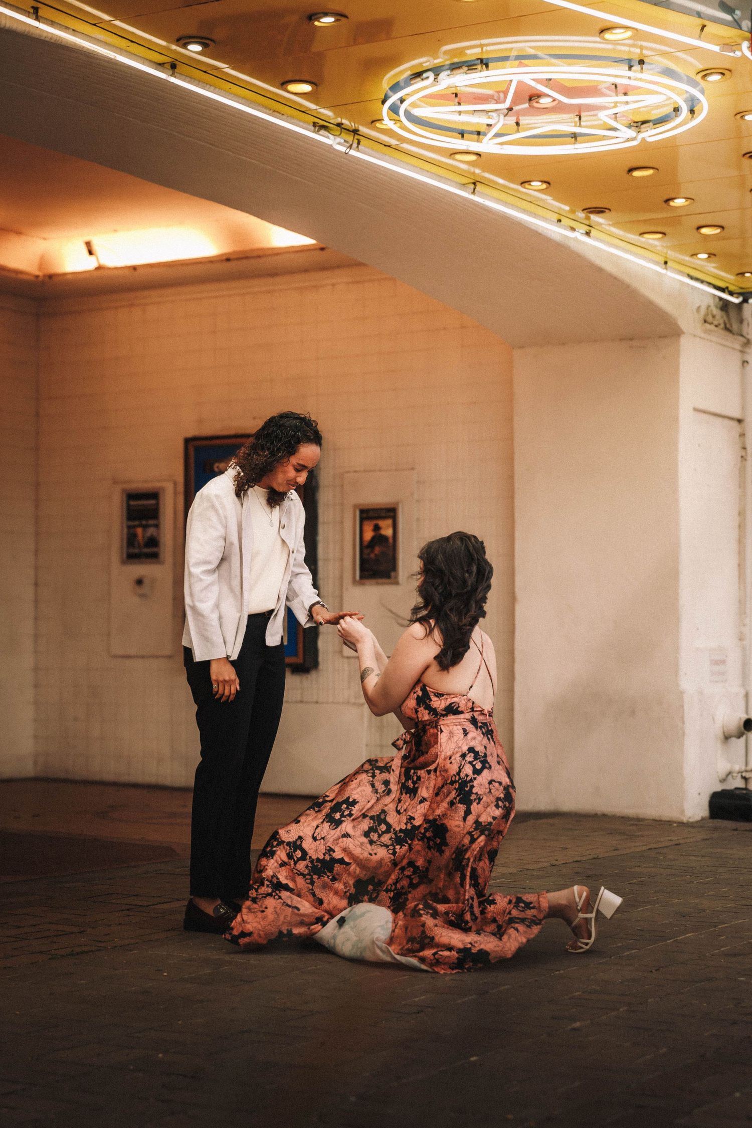 Woman in floral dress kneels proposing to partner in white shirt inside a theater.