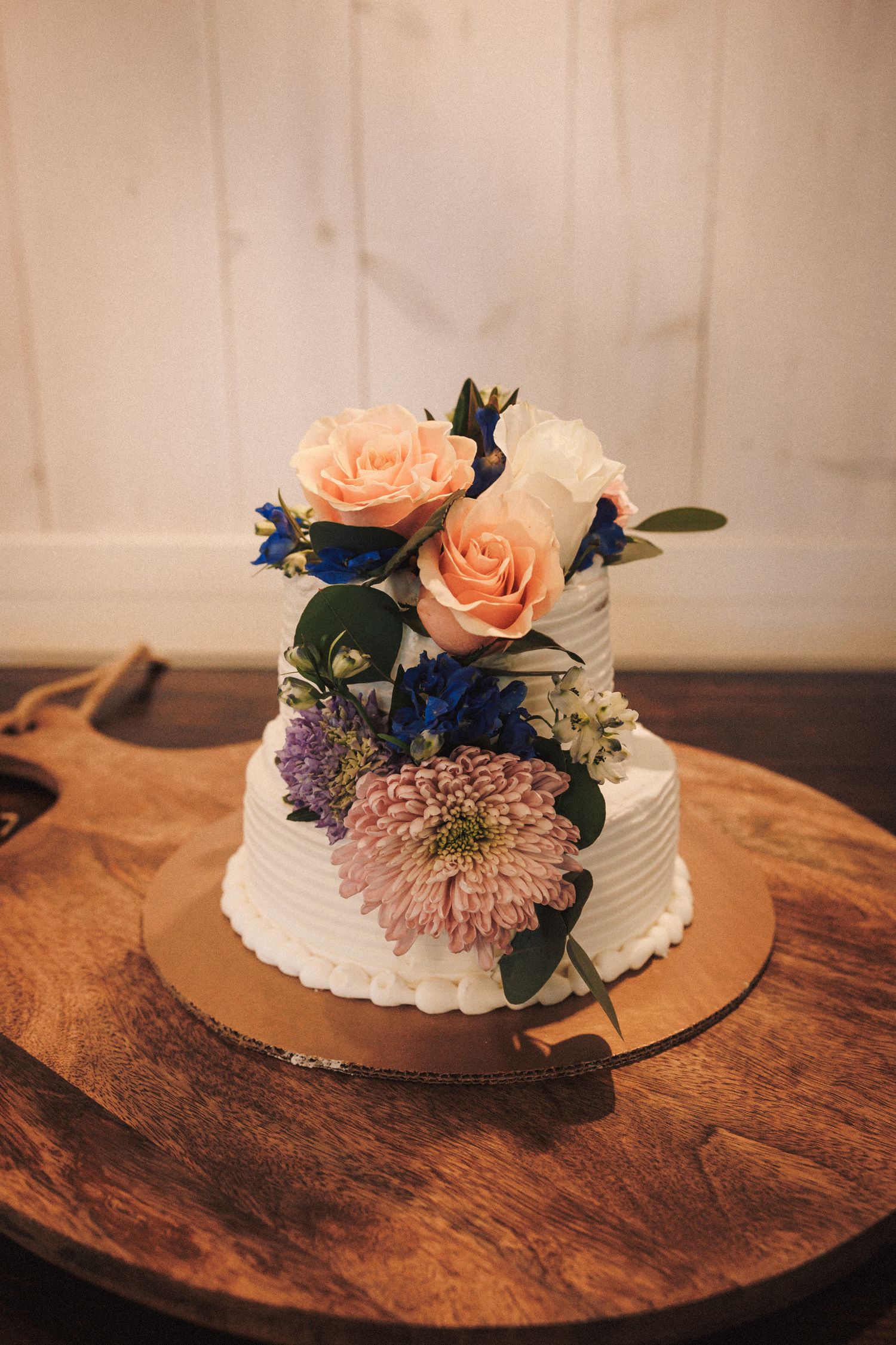 Two-tier white wedding cake adorned with peach roses, purple chrysanthemums, and blue flowers on a wooden cake board.