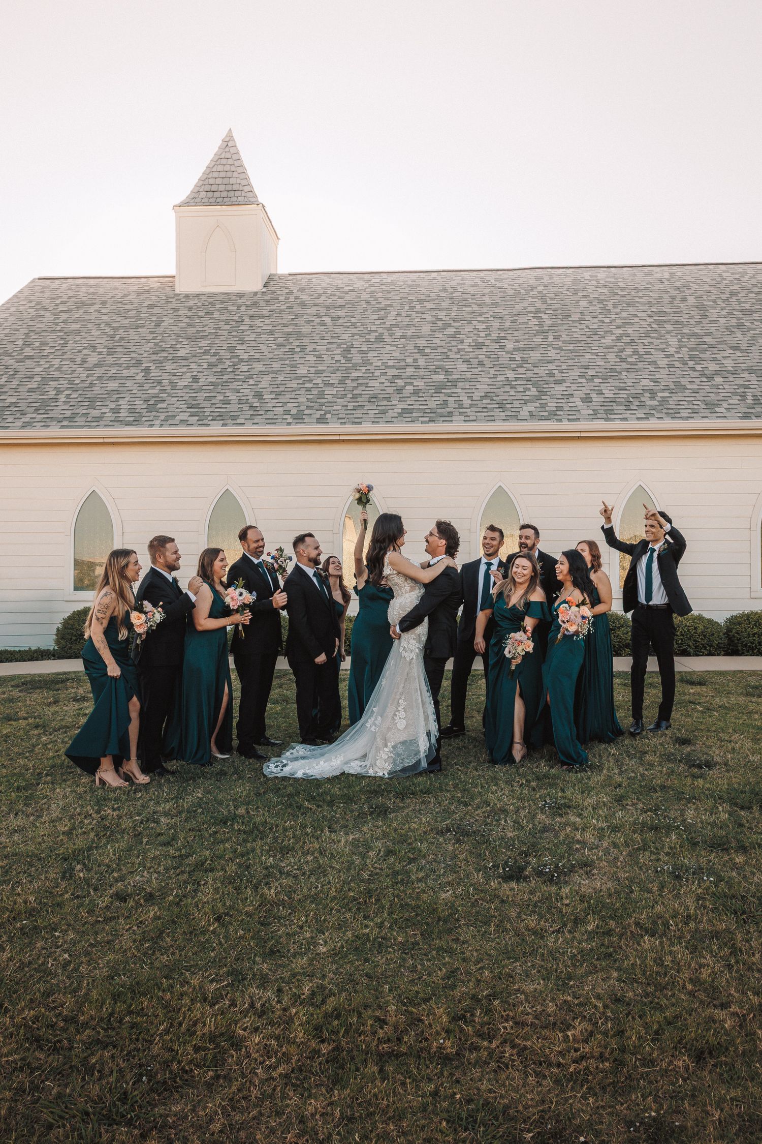 Wedding party in emerald green dresses and black suits celebrating outside white chapel at sunset.