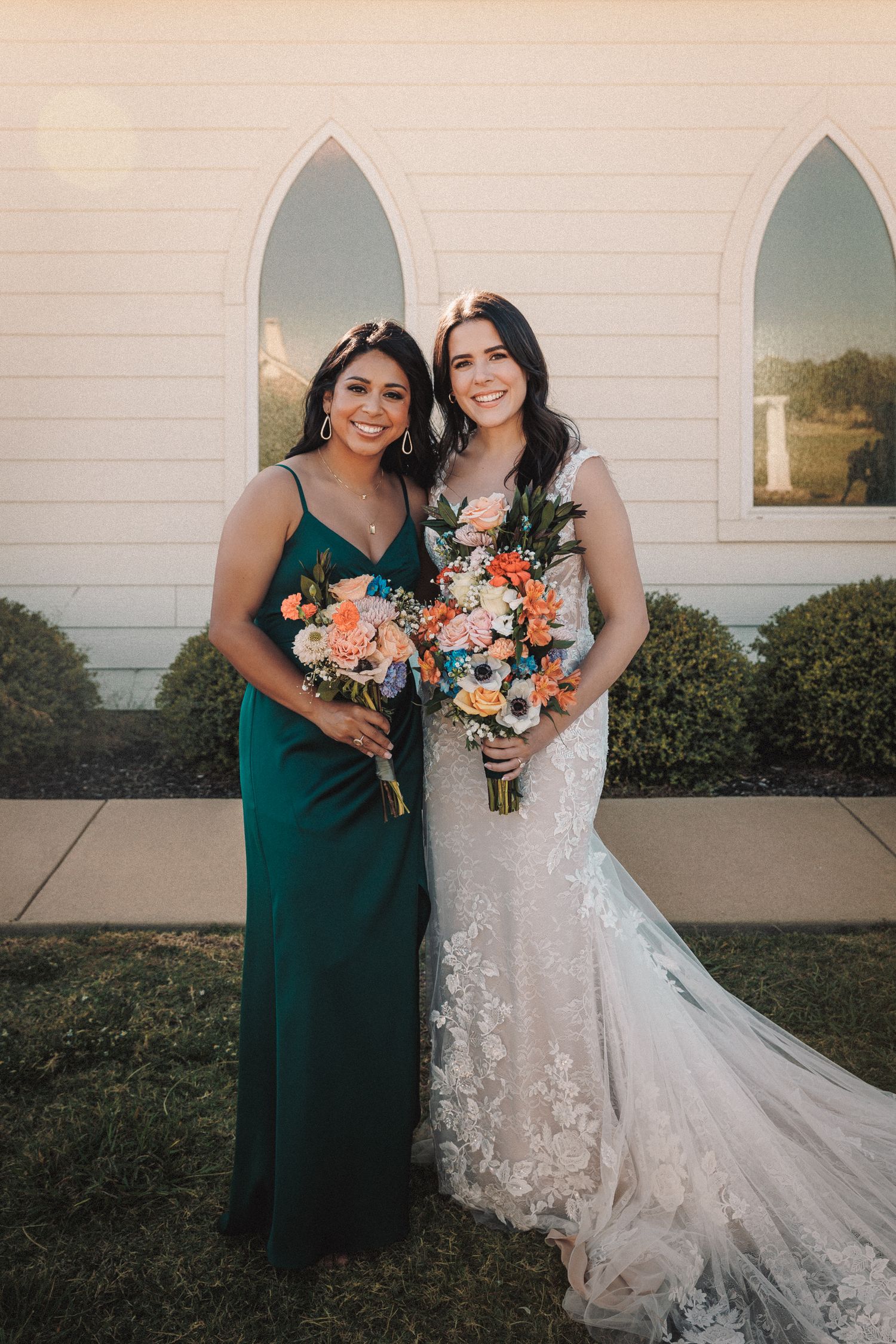 Bride in lace gown and bridesmaid in green dress holding bouquets outside a chapel.