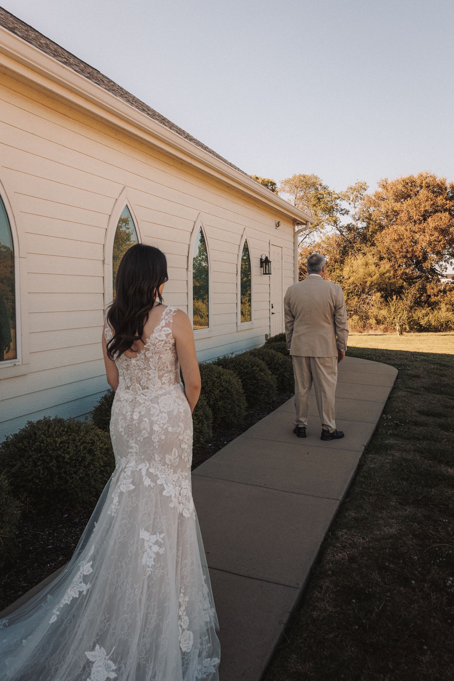 Bride in lace mermaid gown approaches a white chapel for a first look with her father on a sunny day.