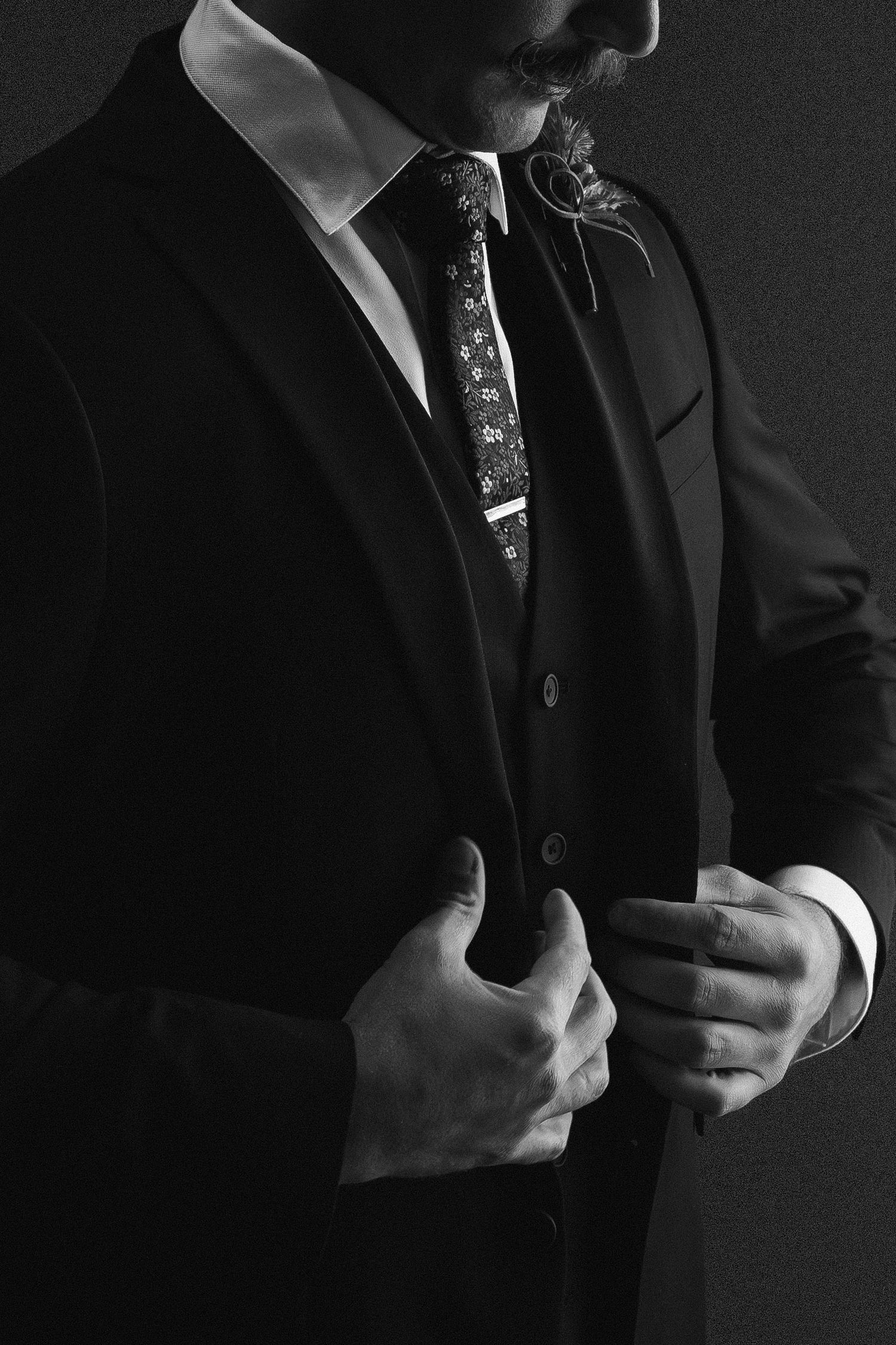 Black and white portrait of a groom in a dark suit adjusting his floral tie.