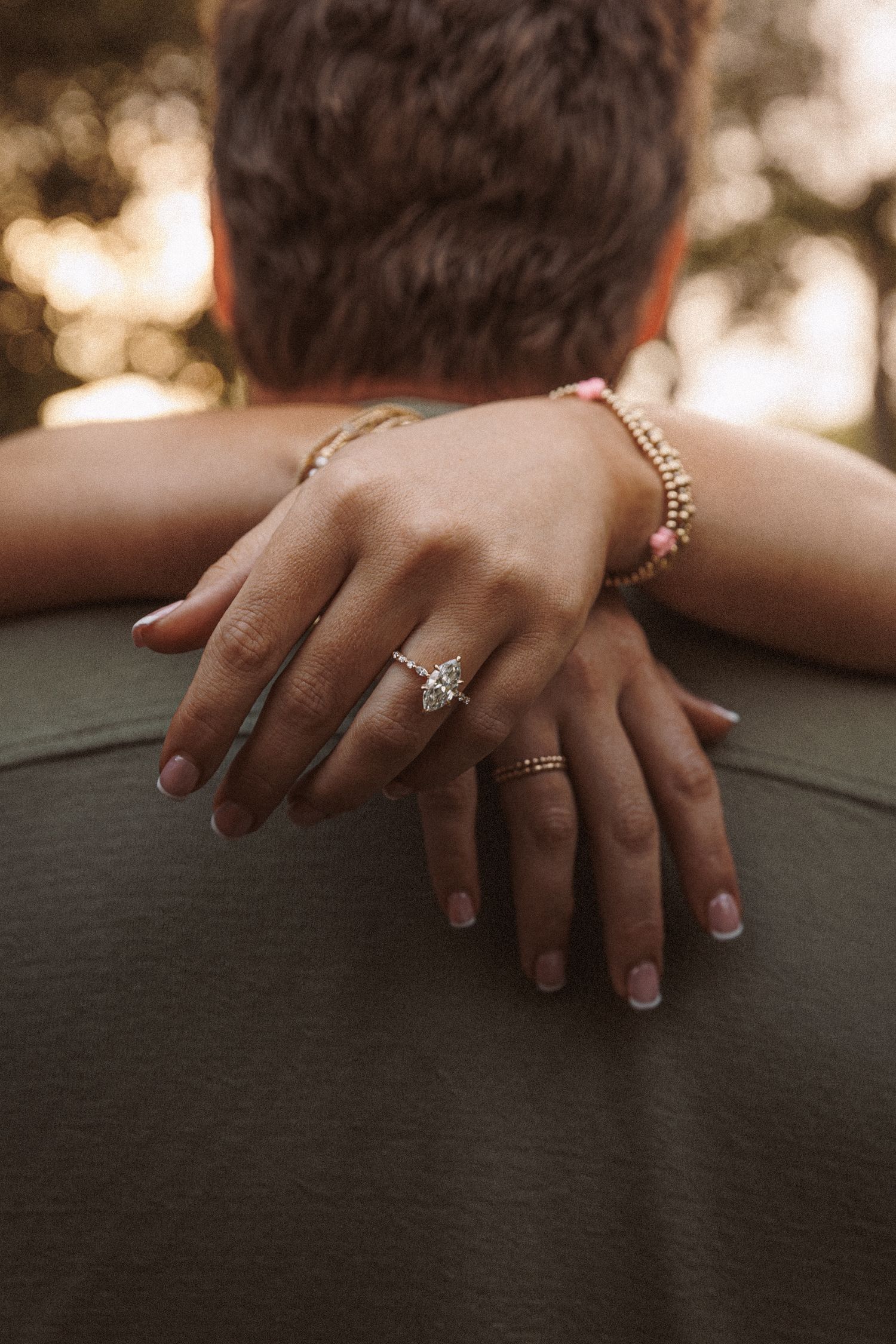 A woman's hand with a diamond engagement ring and beaded bracelets rests on a man's shoulder.