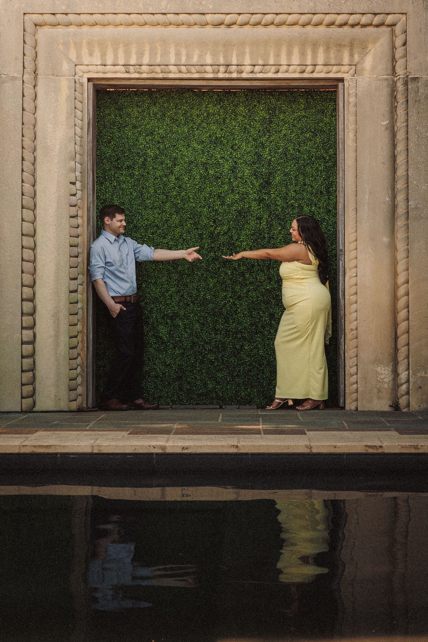 Couple reaching toward each other in front of lush green hedge wall, reflected in pool below.