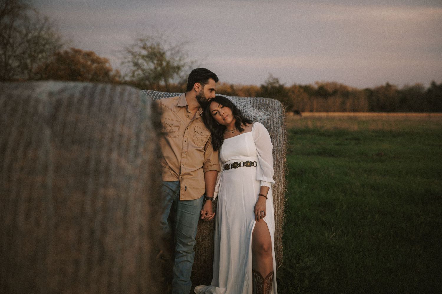 Couple posing romantically by a hay bale in a field at sunset, woman in white dress, man in tan shirt.