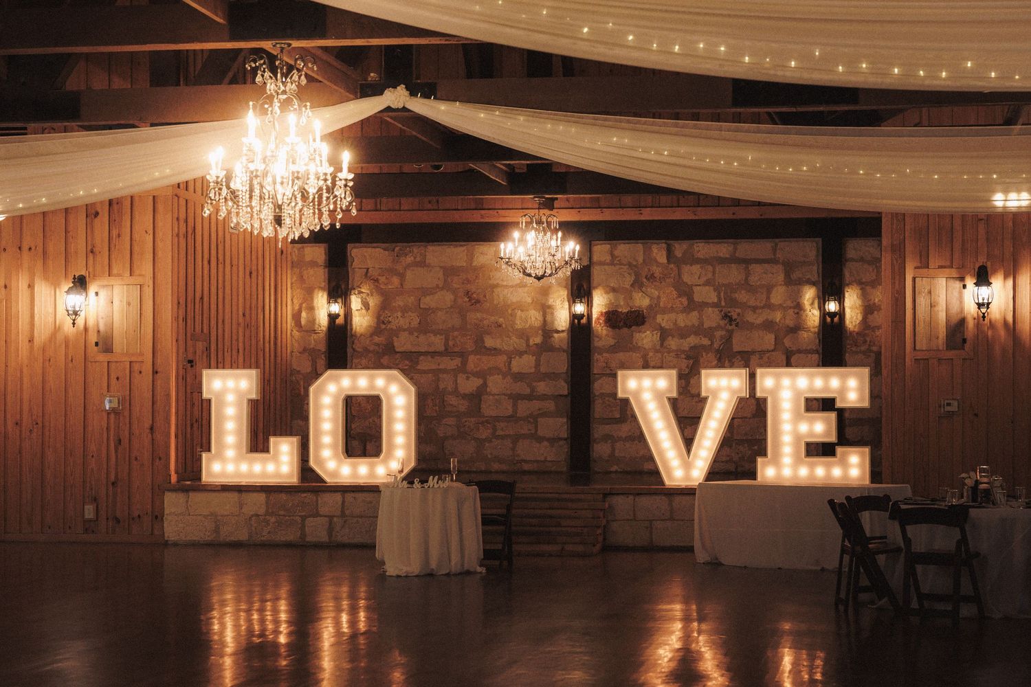 Rustic wedding venue with illuminated LOVE marquee letters, crystal chandeliers, and draped ceiling decor.