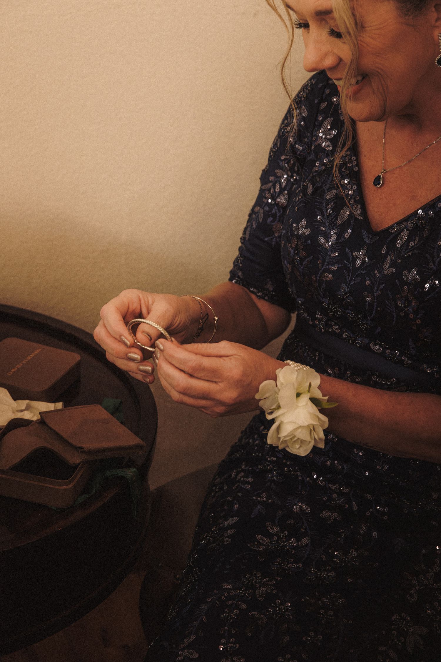 Smiling woman in navy sequin dress with white wrist corsage handles wedding rings at reception.