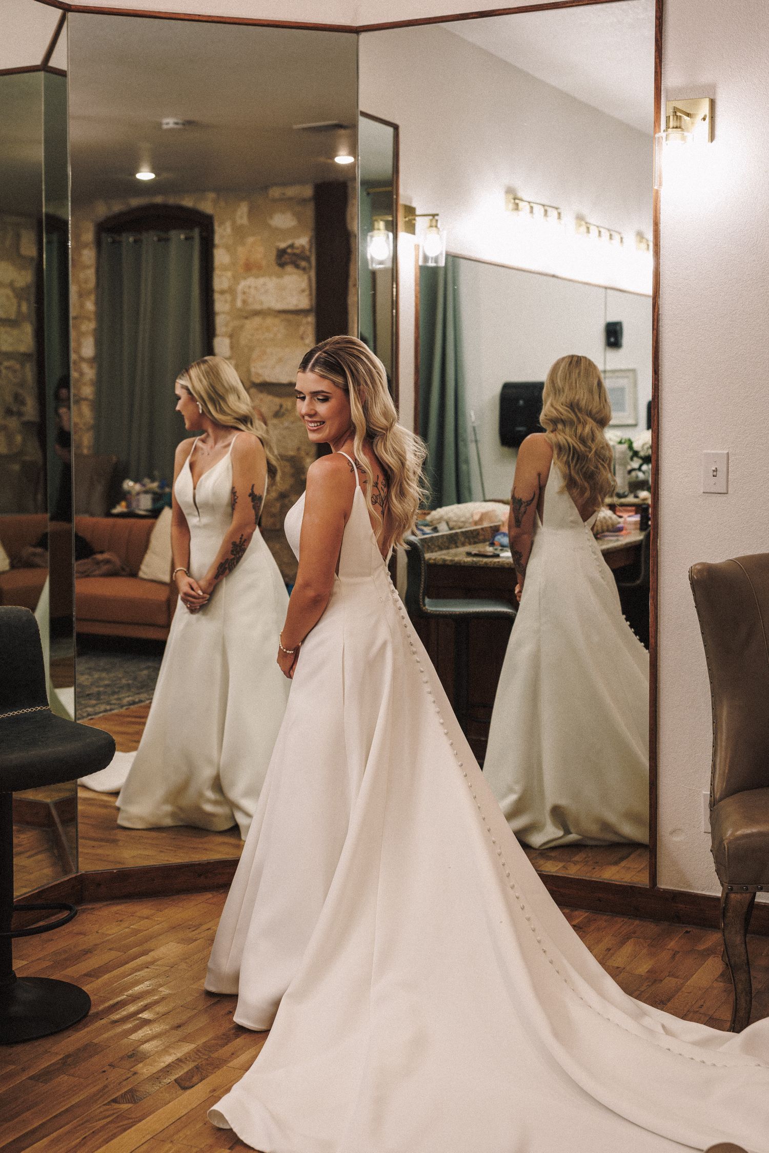 Smiling bride in elegant white satin wedding gown with long train posing in front of mirrors.