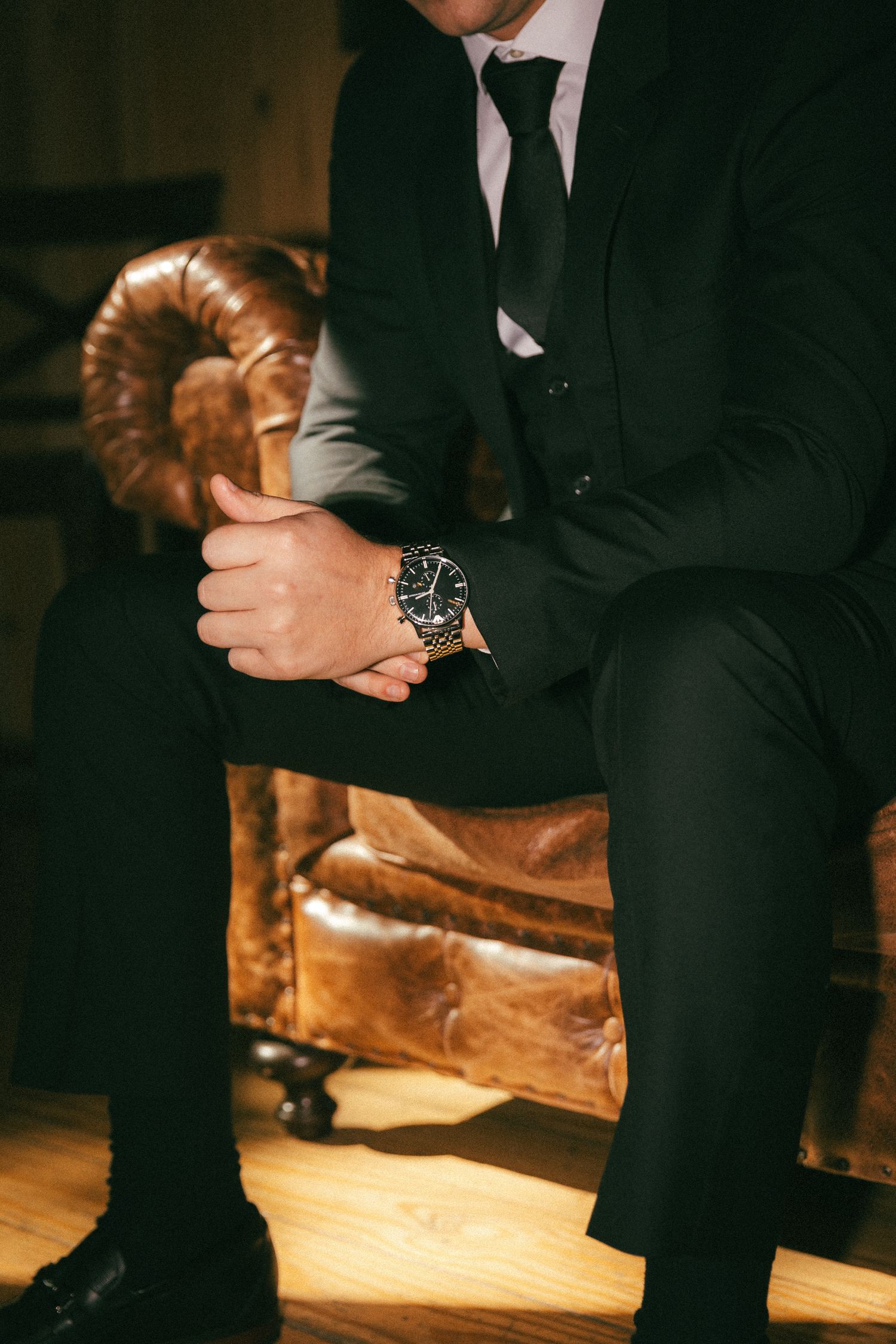 Groom in black suit and tie seated in leather chair adjusting his watch before the wedding ceremony.