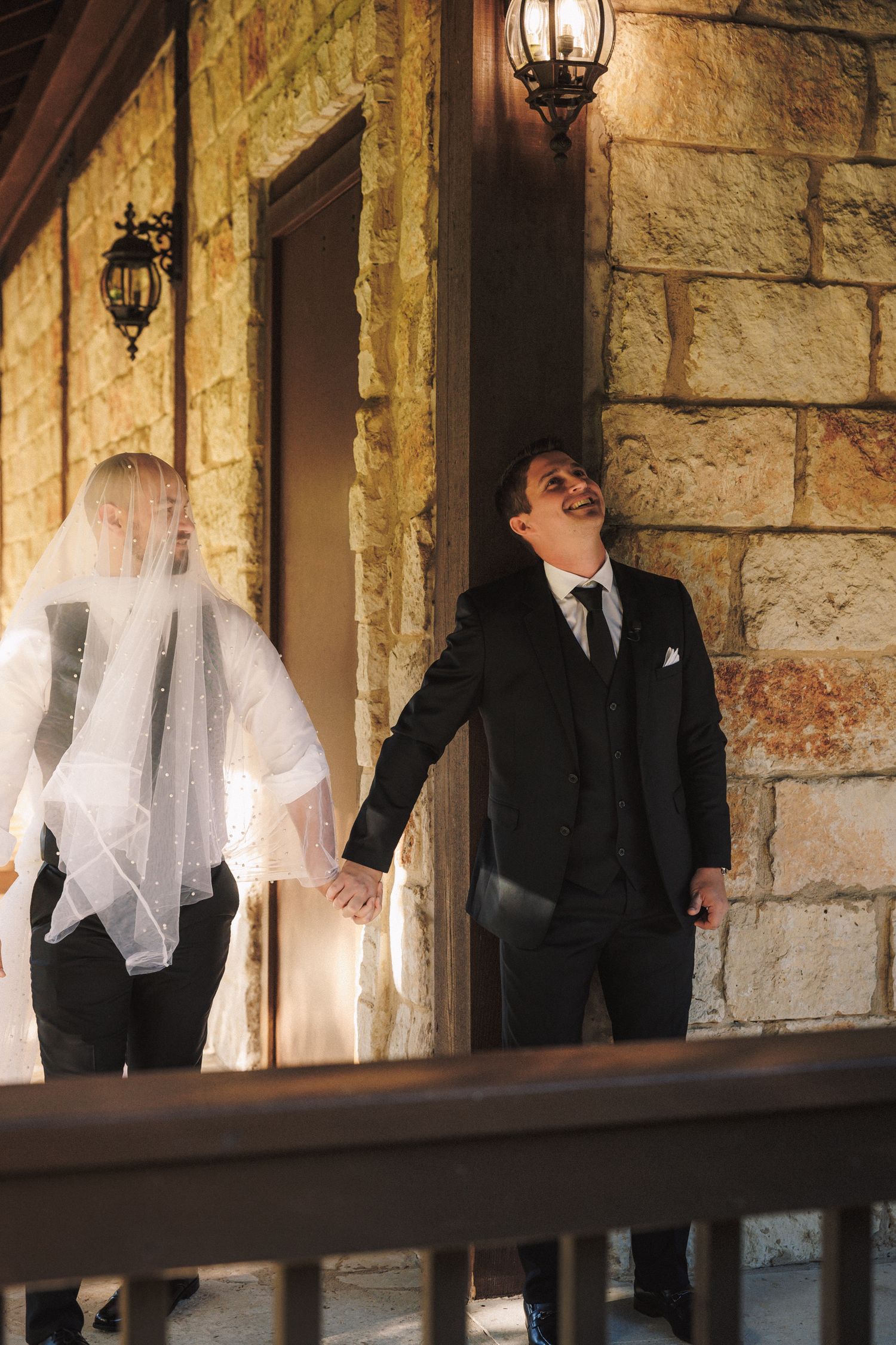 Groom in black suit laughing joyfully while holding hands with bride in veil outside stone building.