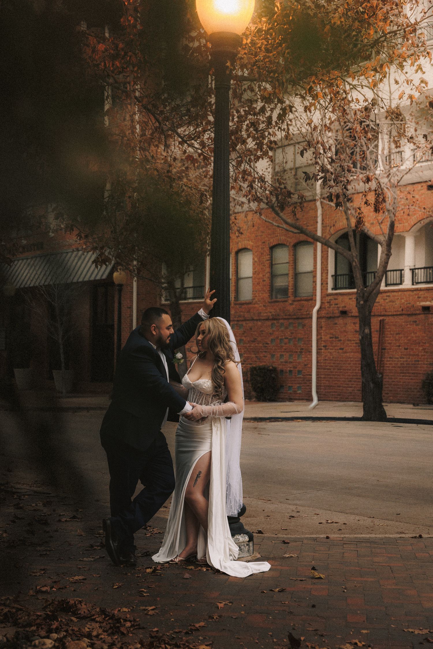 A romantic couple shares an intimate moment at sunset in front of brick buildings with autumn leaves scattered around.