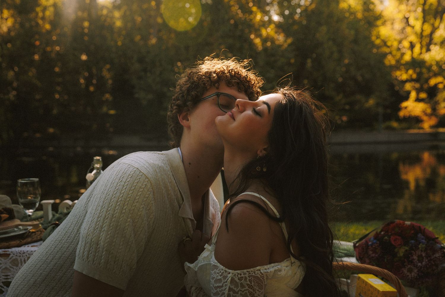 A couple shares a romantic kiss during golden hour in a scenic outdoor setting with sunlit trees in the background.