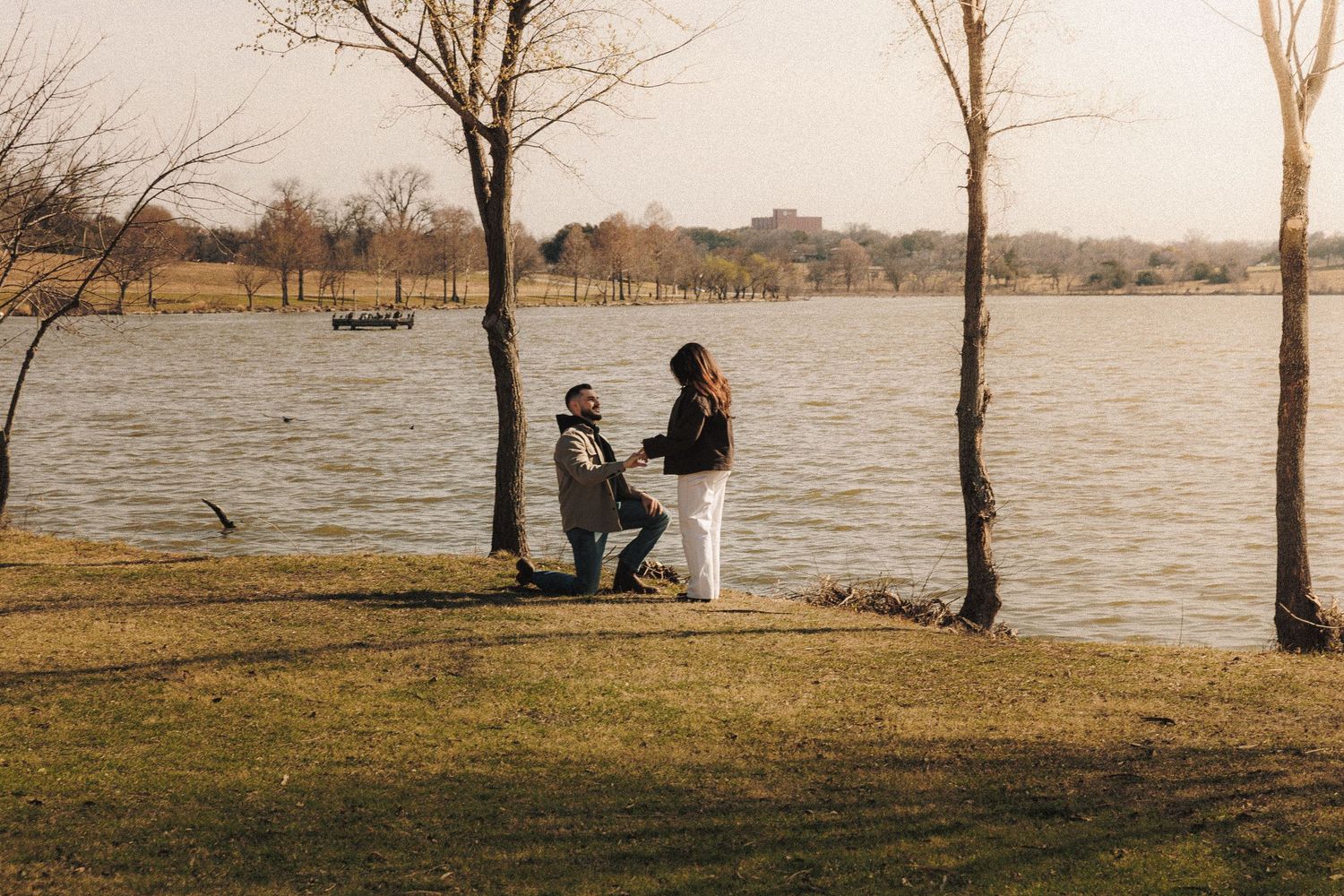 A romantic lakeside proposal scene at sunset with silhouettes against the water.